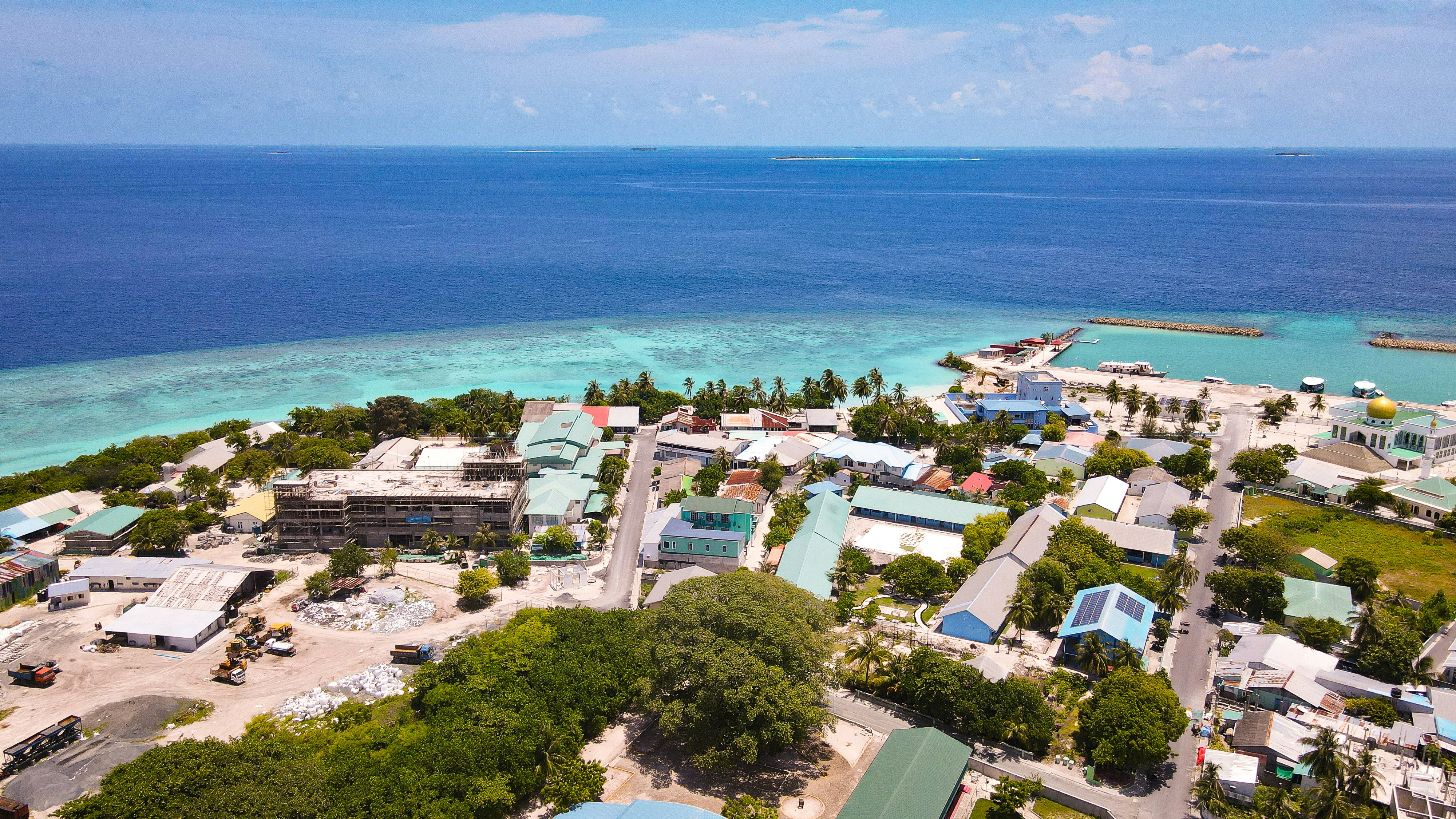 An aerial view of Ungoofaaru in the Maldives: houses with colored roofs among palms, a mosque with a gold dome, and a harbor jetty where the reef flat turns from turquoise to deep blue