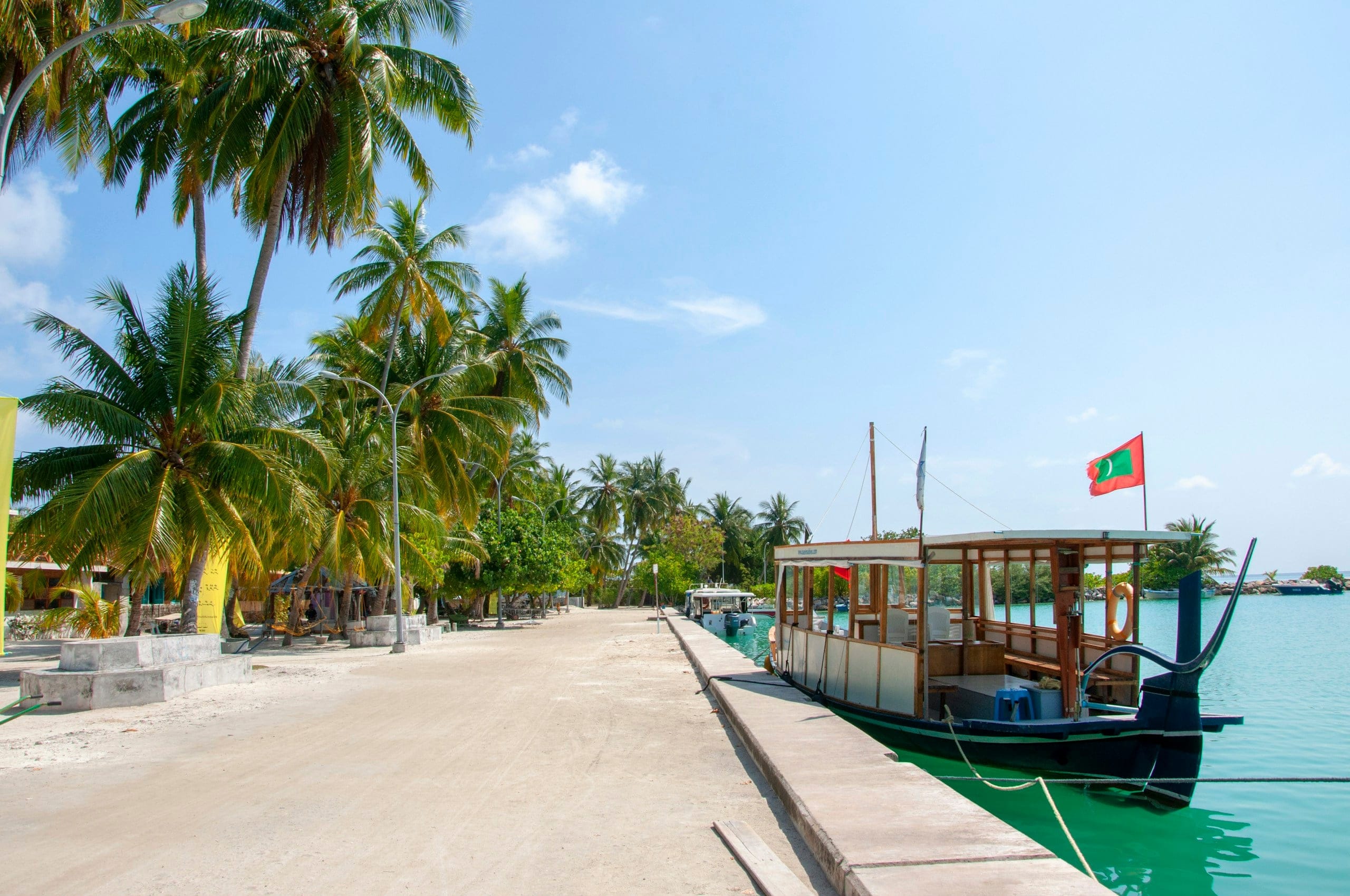 A wooden dhoni tied alongside the concrete jetty at Felidhoo Harbour, the Maldivian flag on its stern and coconut palms lining the shore behind