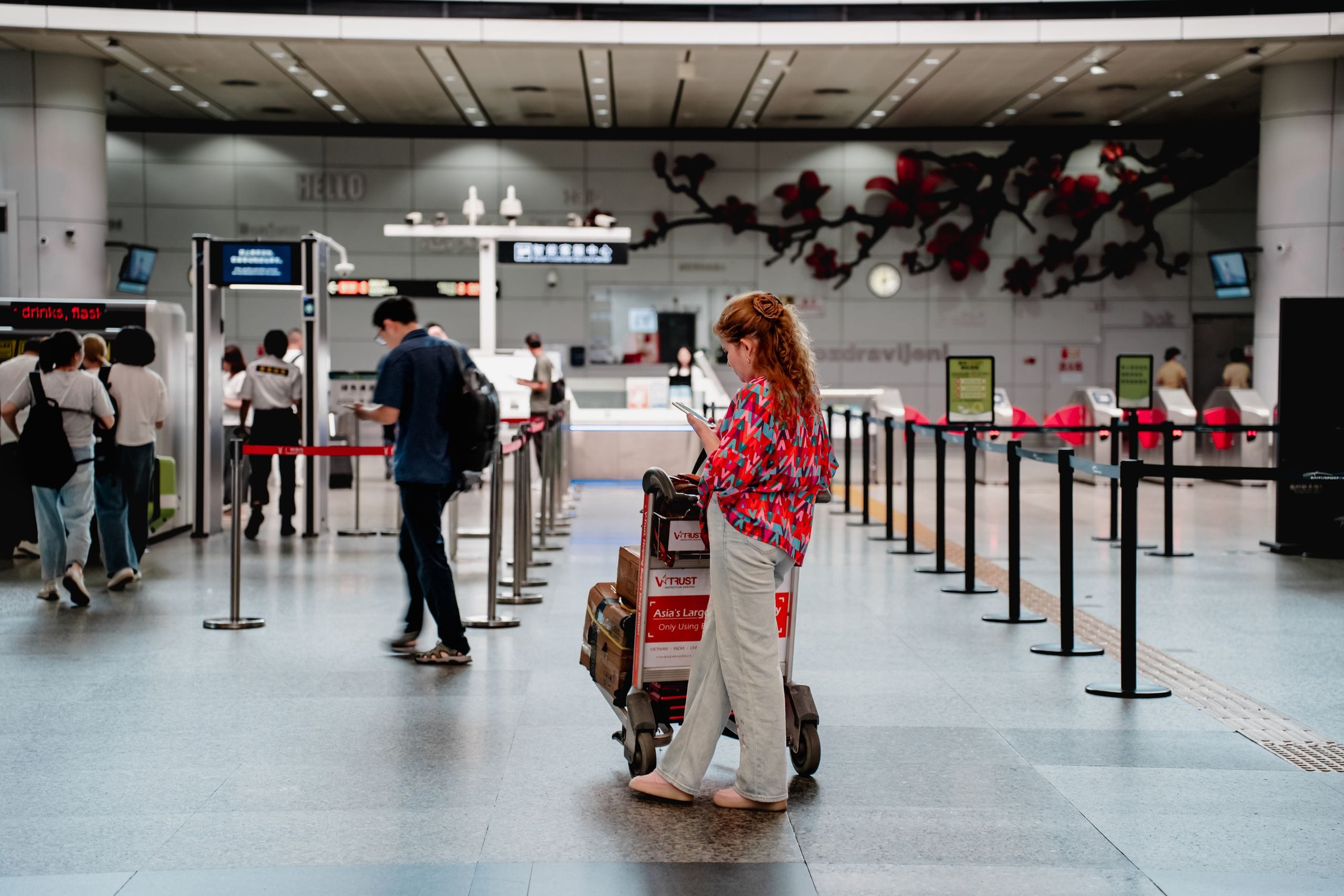 A traveler in Guangzhou checks her phone beside a luggage cart in a terminal hall, other passengers passing behind
