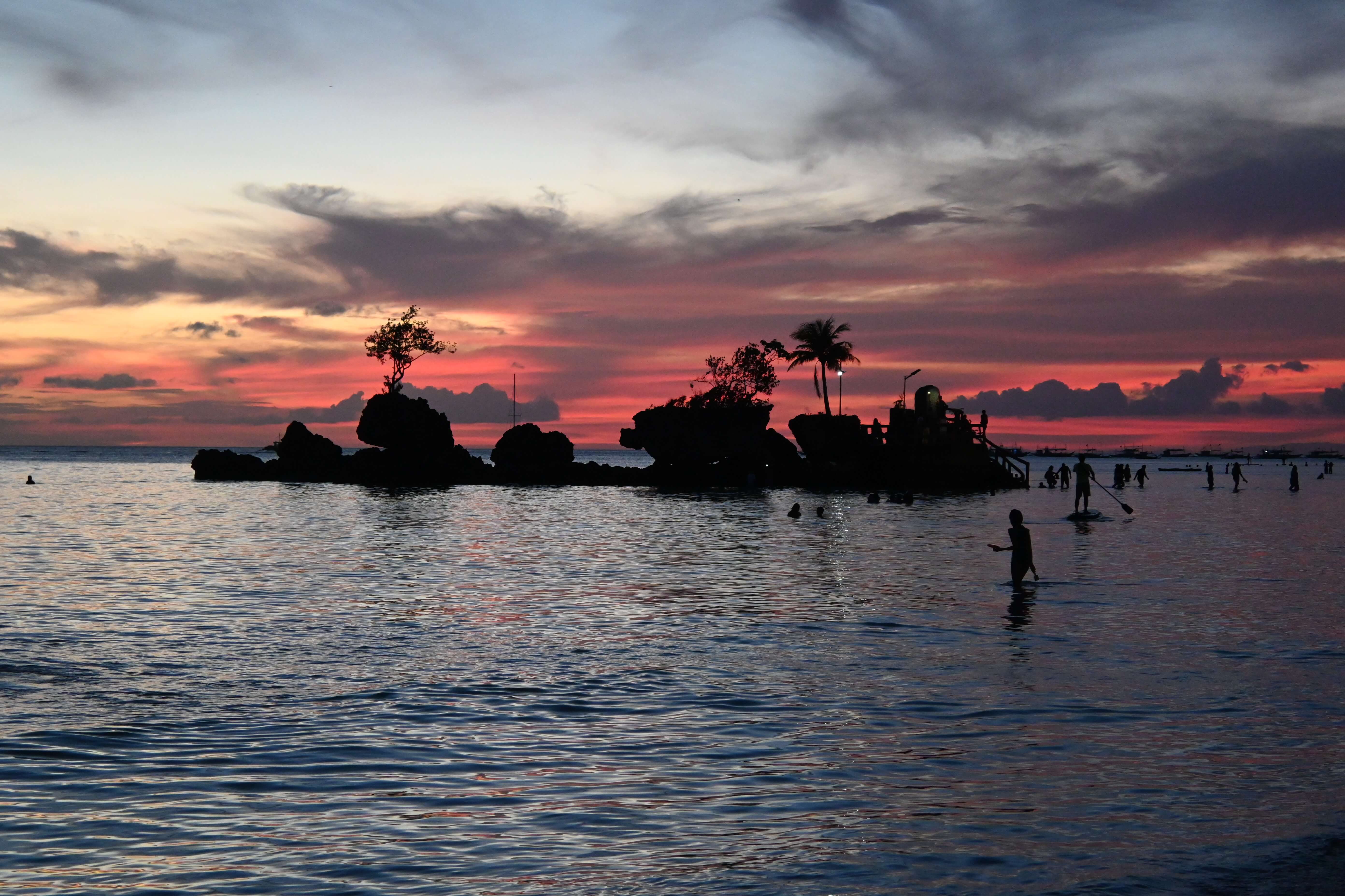 Willy's Rock off White Beach in Boracay, silhouetted at sunset with its shrine steps and a single palm against a red sky, people standing in the shallows