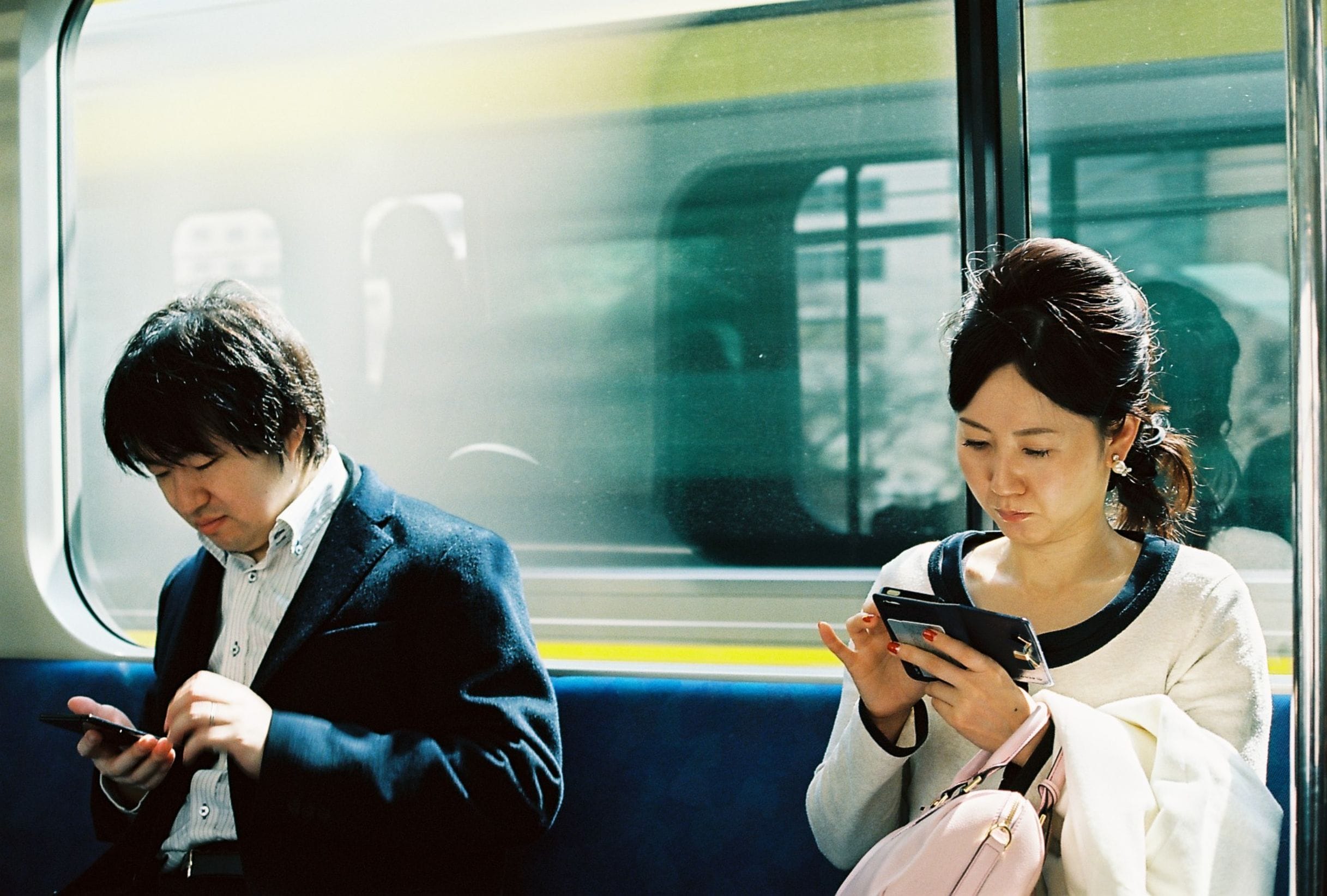 Two commuters on a train, a man in a navy jacket and a woman with a pink bag, both reading their phones