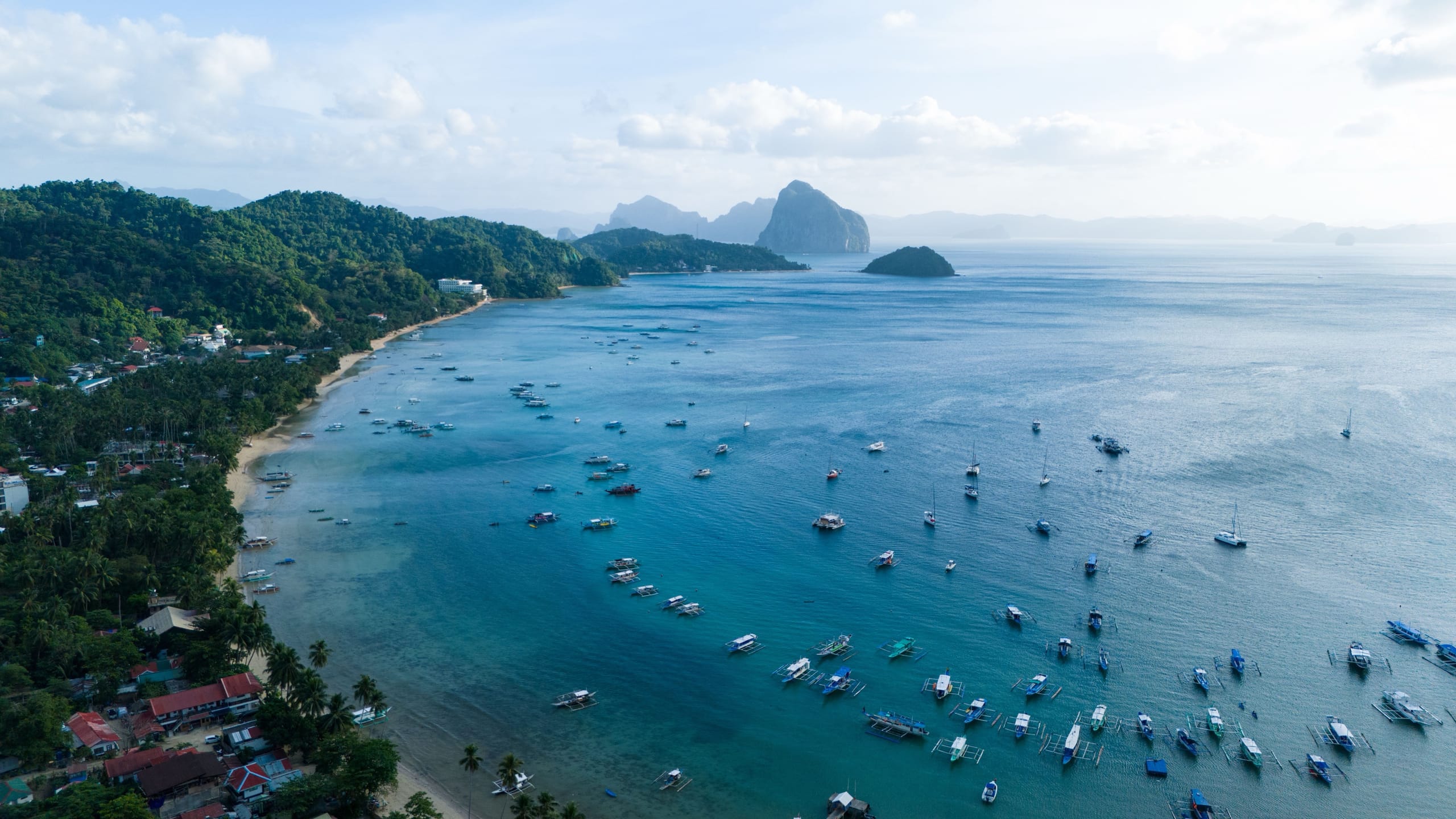 An aerial view of El Nido town beach with outrigger boats moored across the bay and karst islands behind