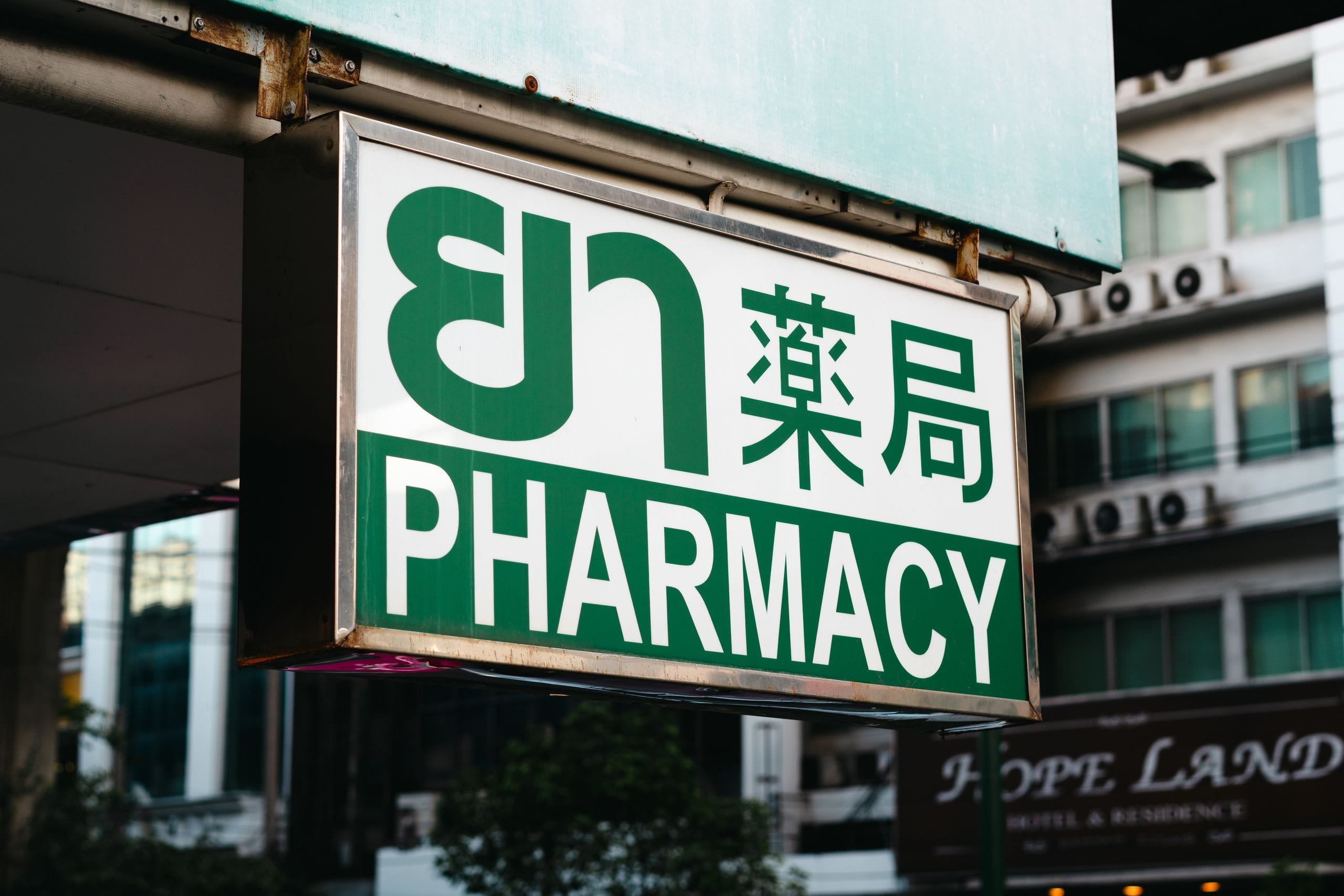 A green and white pharmacy sign in English, Thai and Chinese hanging over a city street