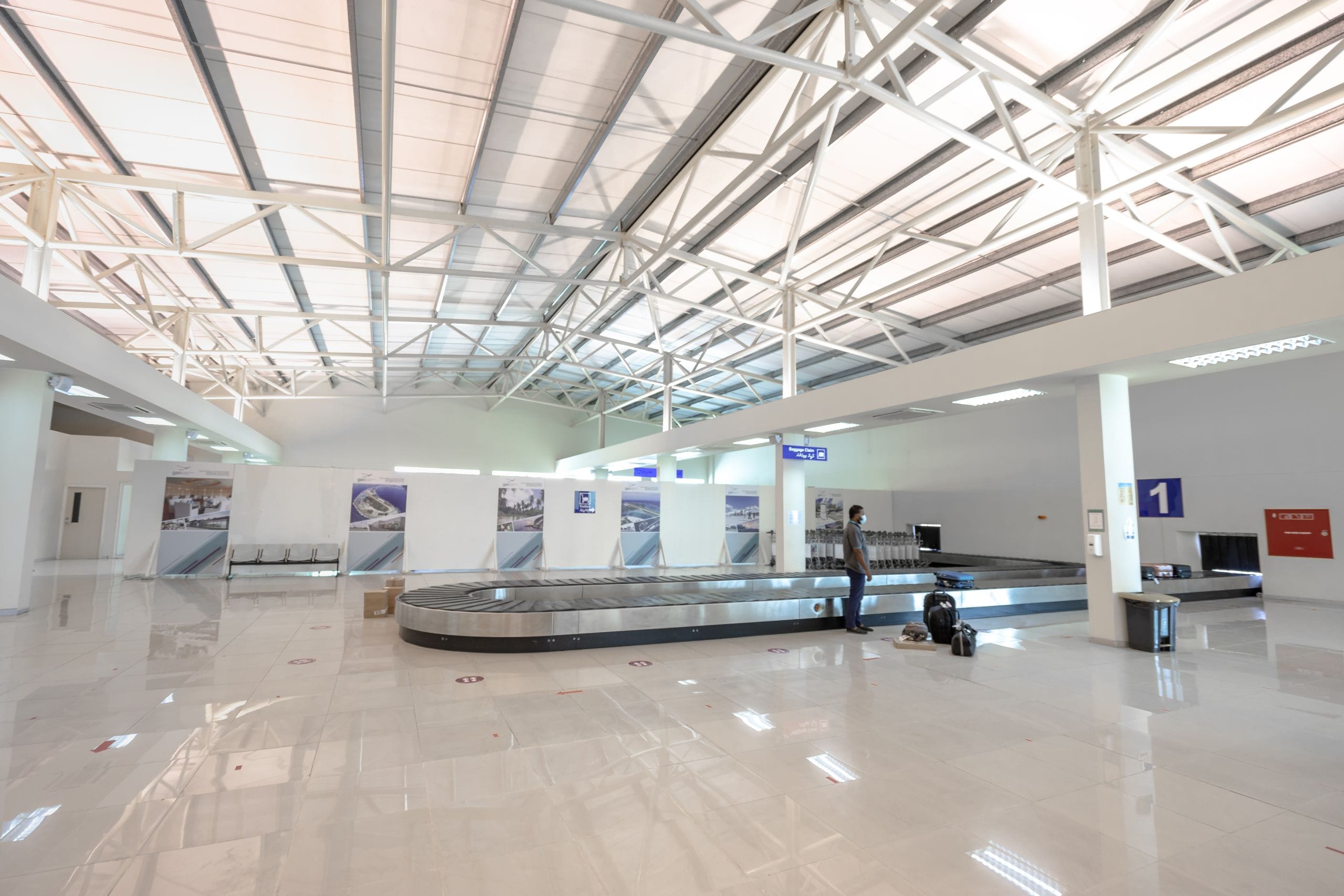 A bright baggage claim hall with one traveler waiting beside the carousel and a few bags
