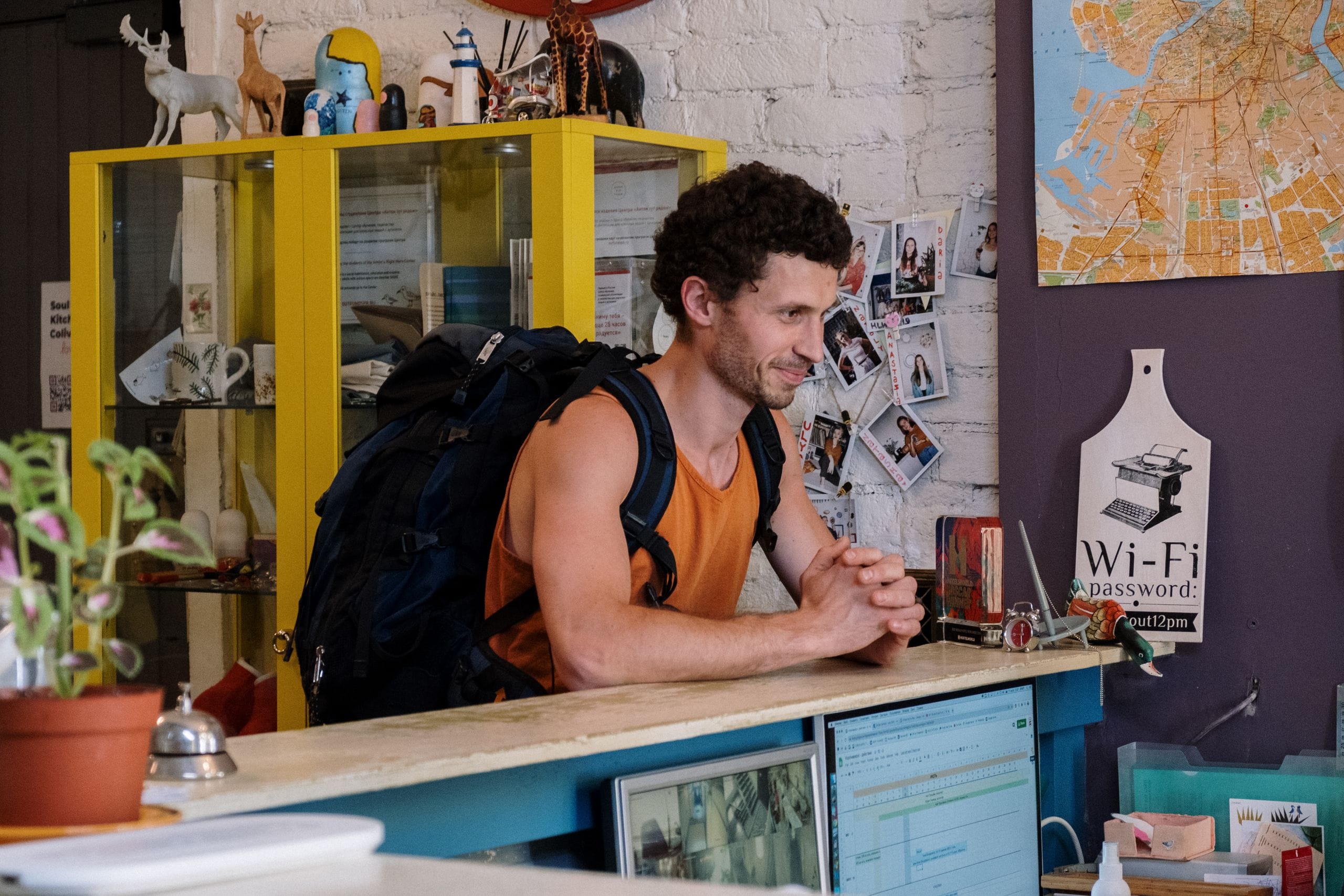 A backpacker leaning on a hostel reception desk, a city map and a wifi password sign on the wall behind