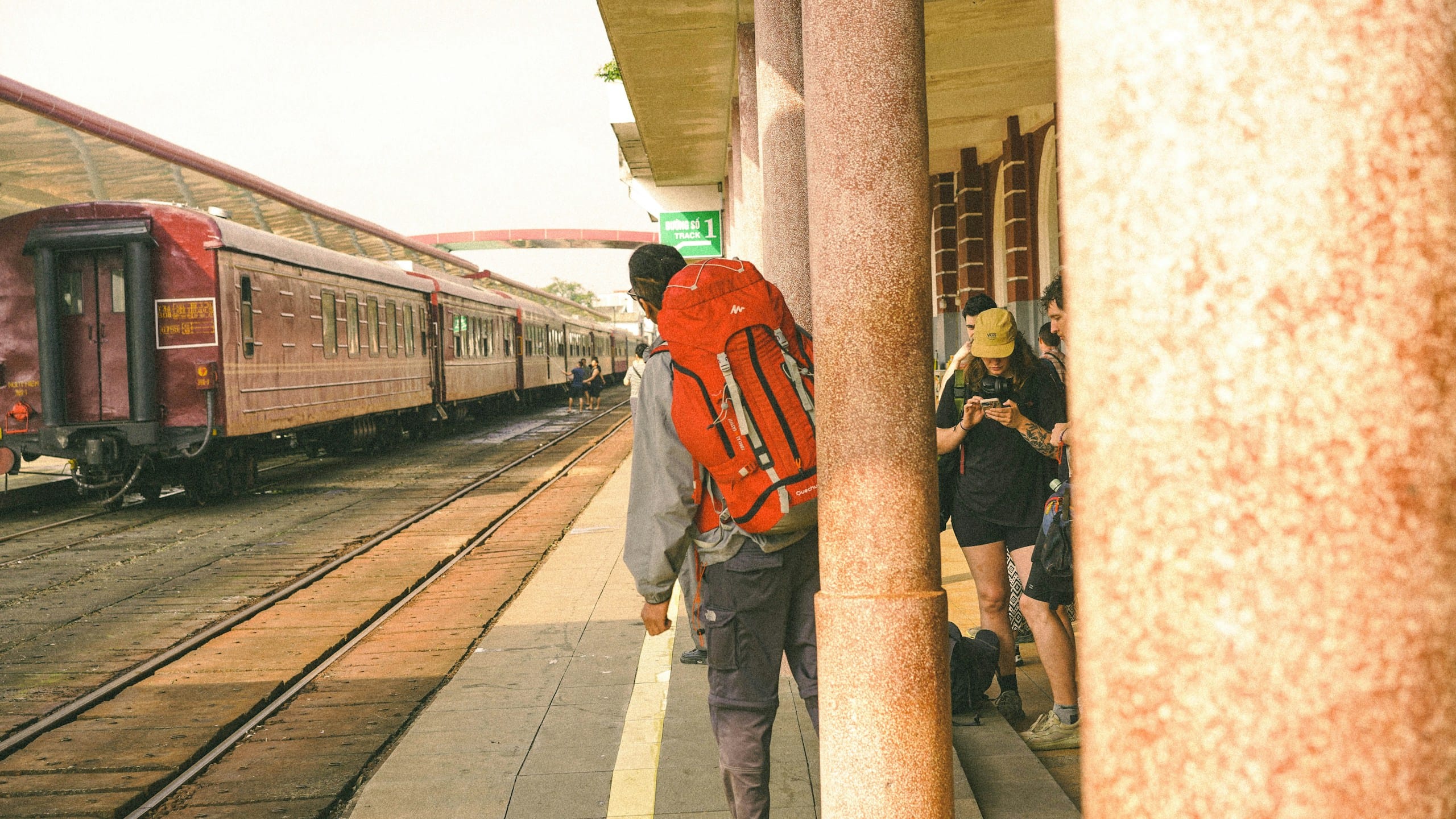 A man with a big red backpack on a station platform beside a row of old maroon passenger carriages