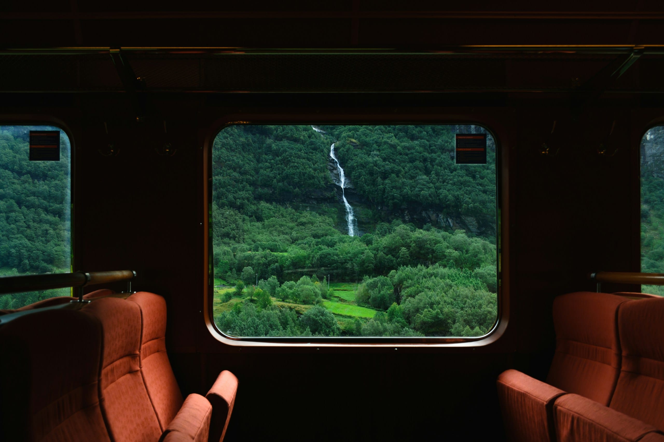 A waterfall on a green forested mountainside, framed by a train window between empty orange seats