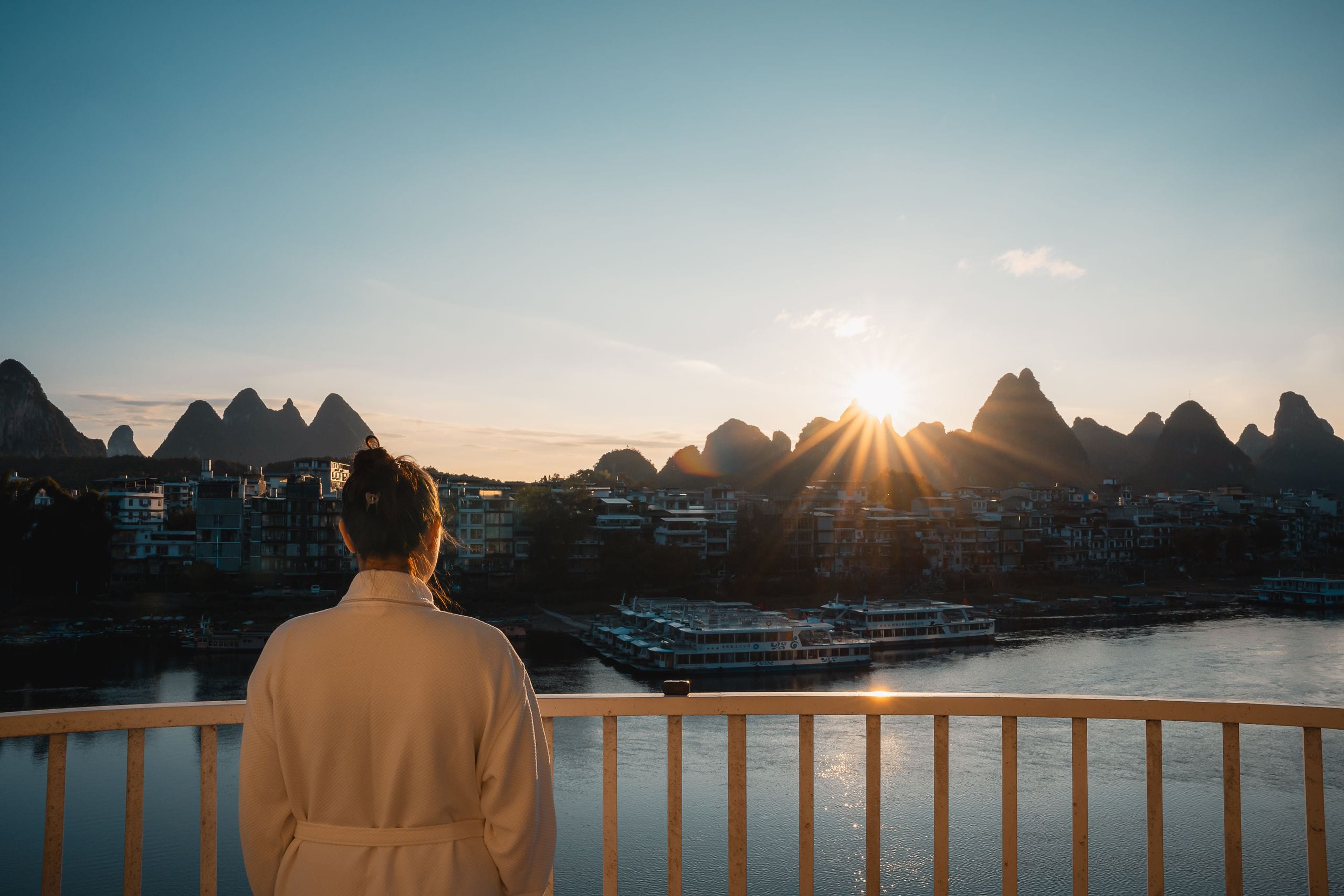 A woman in a robe on a balcony at sunrise, facing the karst peaks and river boats of Guilin