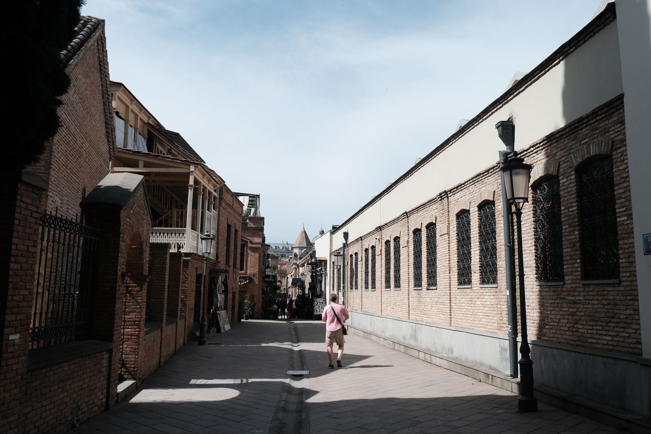 A man in a pink shirt walking a sunny brick street in the Tbilisi old town, wooden balconies on the left