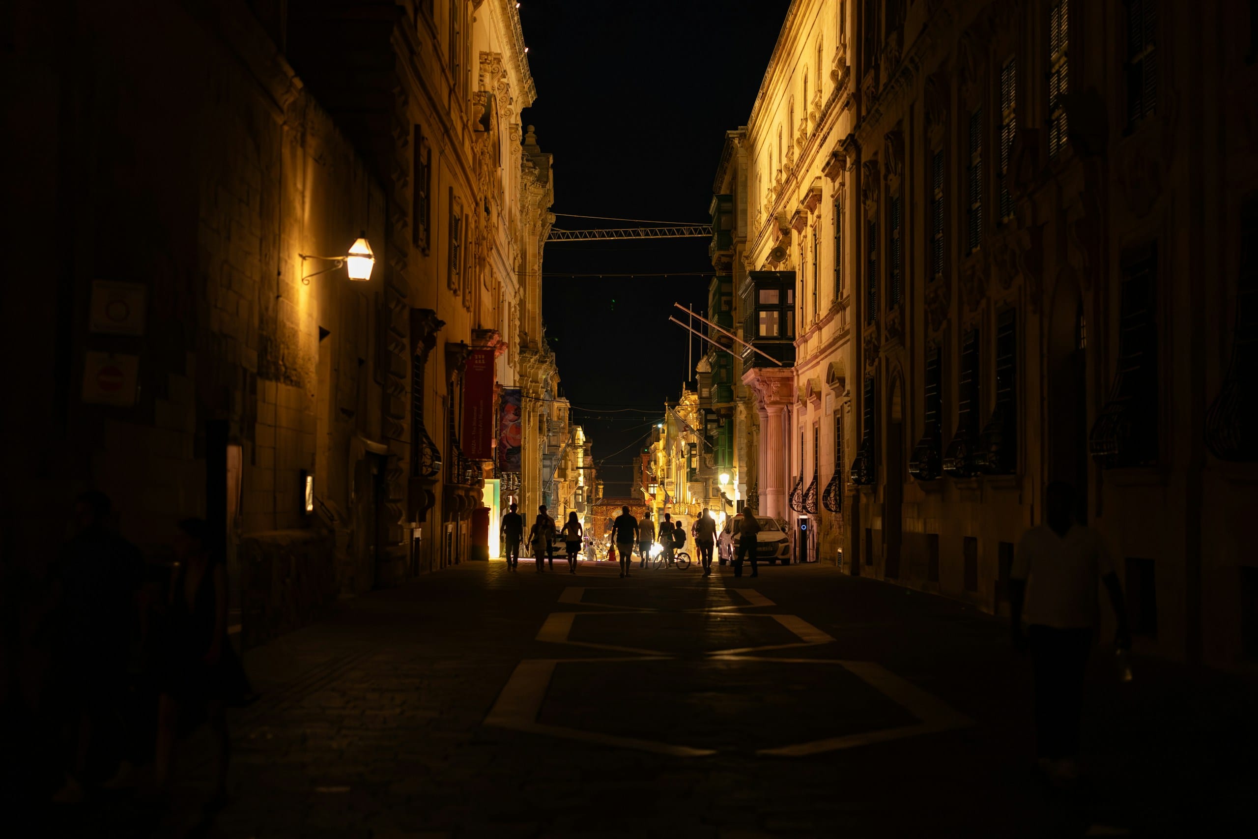 A narrow stone street at night lit by warm lamps, a few people walking toward the bright far end