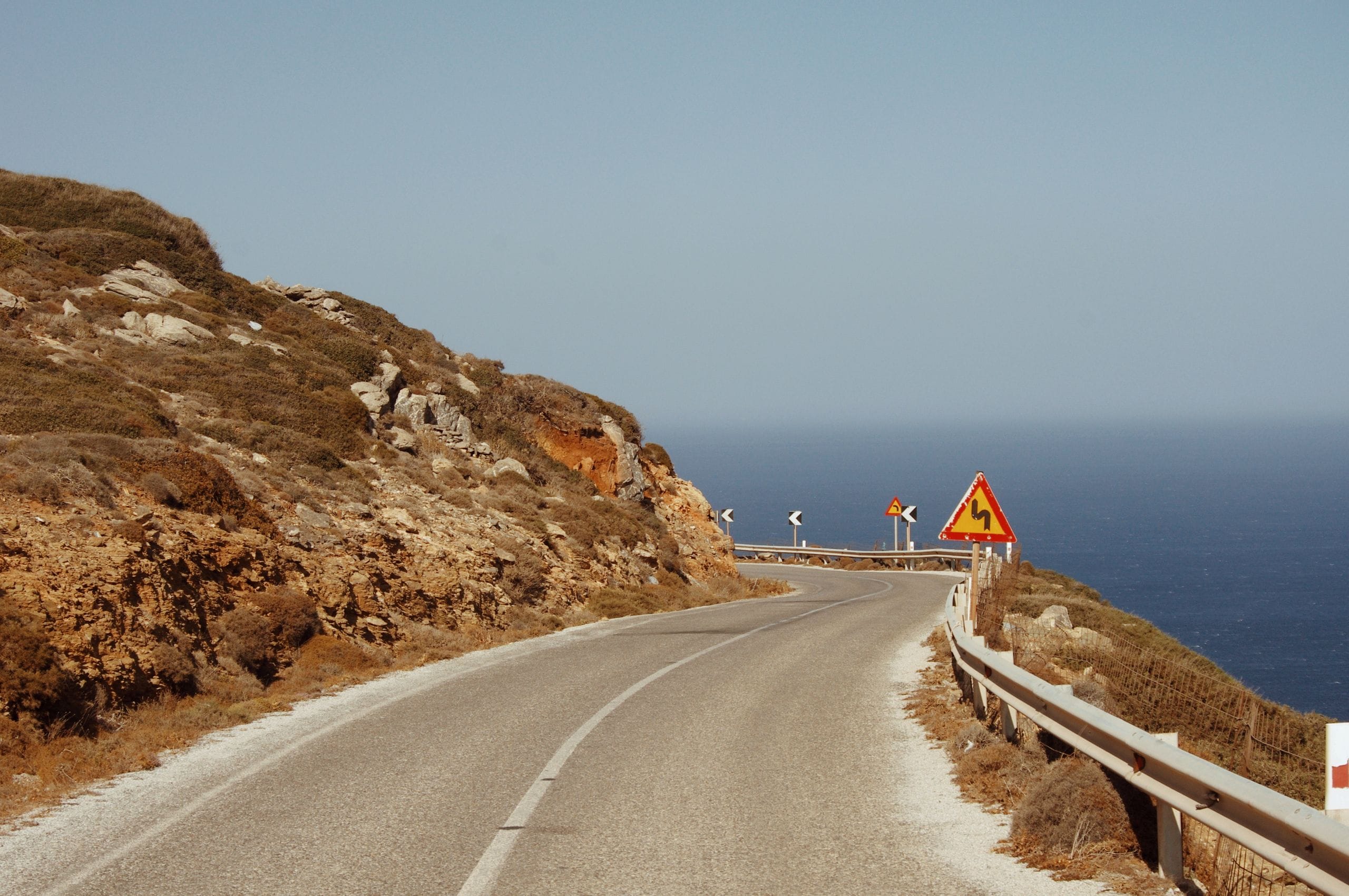 A two-lane road curving along a dry hillside above the sea, with a winding-road warning sign and guardrail
