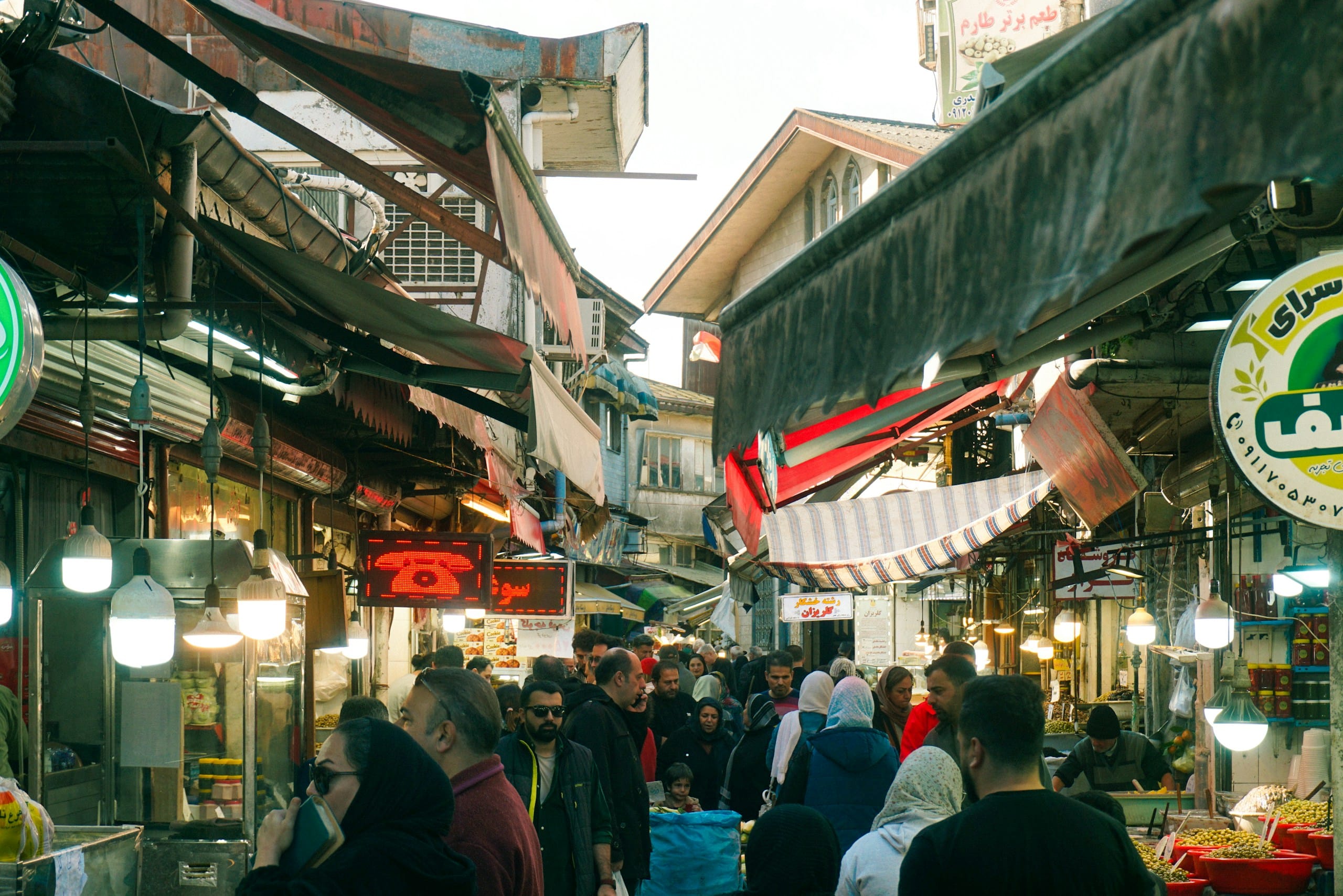Shoppers packed shoulder to shoulder in a covered market lane under striped awnings and hanging lamps