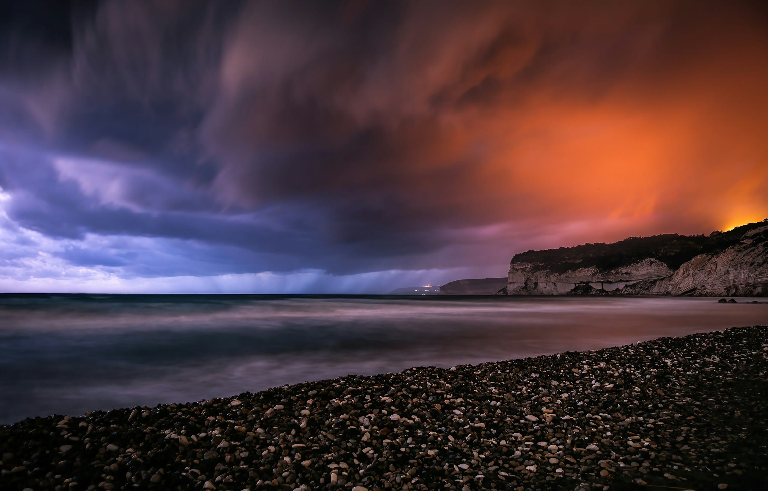Storm clouds lit orange and violet rolling over the sea, a pebble beach and pale cliffs in the foreground