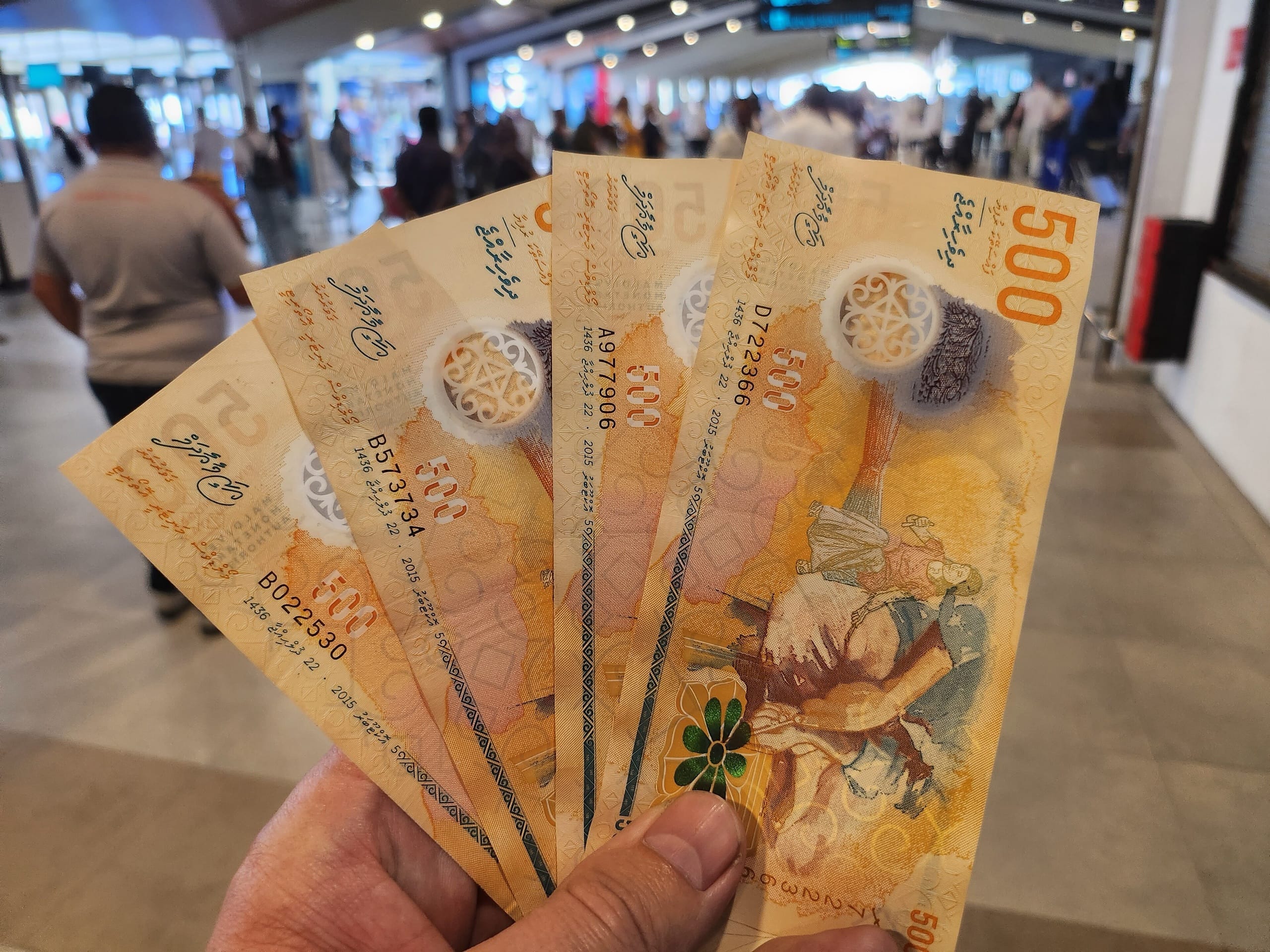 A hand holding a fan of orange 500 rufiyaa banknotes in a busy terminal hall