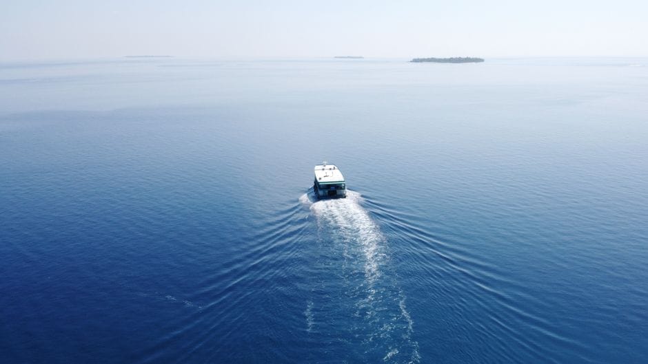 A small passenger boat crossing open water in the Maldives, its wake trailing behind it, with a low green island on the far horizon.