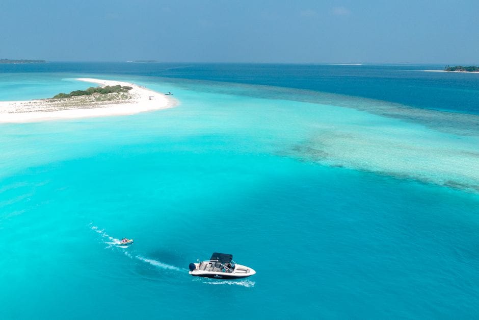 A speedboat crossing pale turquoise shallows in the Maldives toward a low sandy island, with the water turning dark blue where the reef drops away.