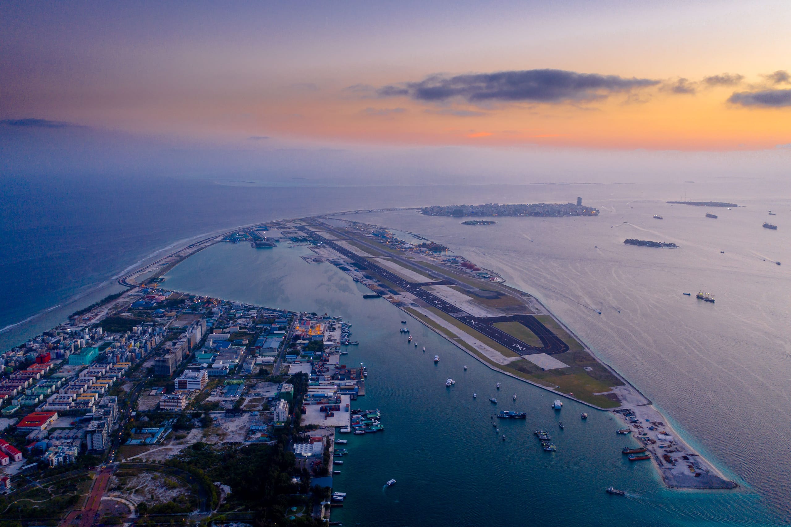 An aerial view at dusk of the airport island at Male: a single runway on a strip of reclaimed land, a channel of moored boats between it and a dense island of low-rise blocks, and open sea in every direction.
