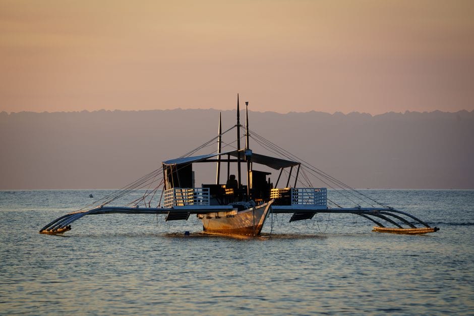 A Filipino outrigger boat with long bamboo arms sitting on flat water at first light in Central Visayas, a low hill line behind it
