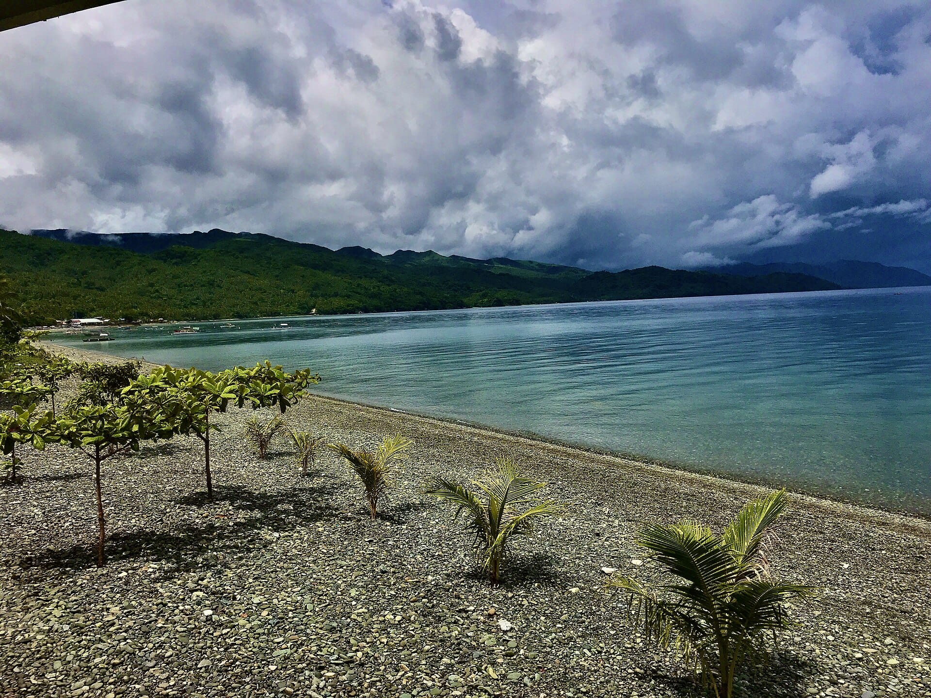 Dark storm clouds building over green hills and a calm gulf, seen from a pebble beach in Camarines Sur