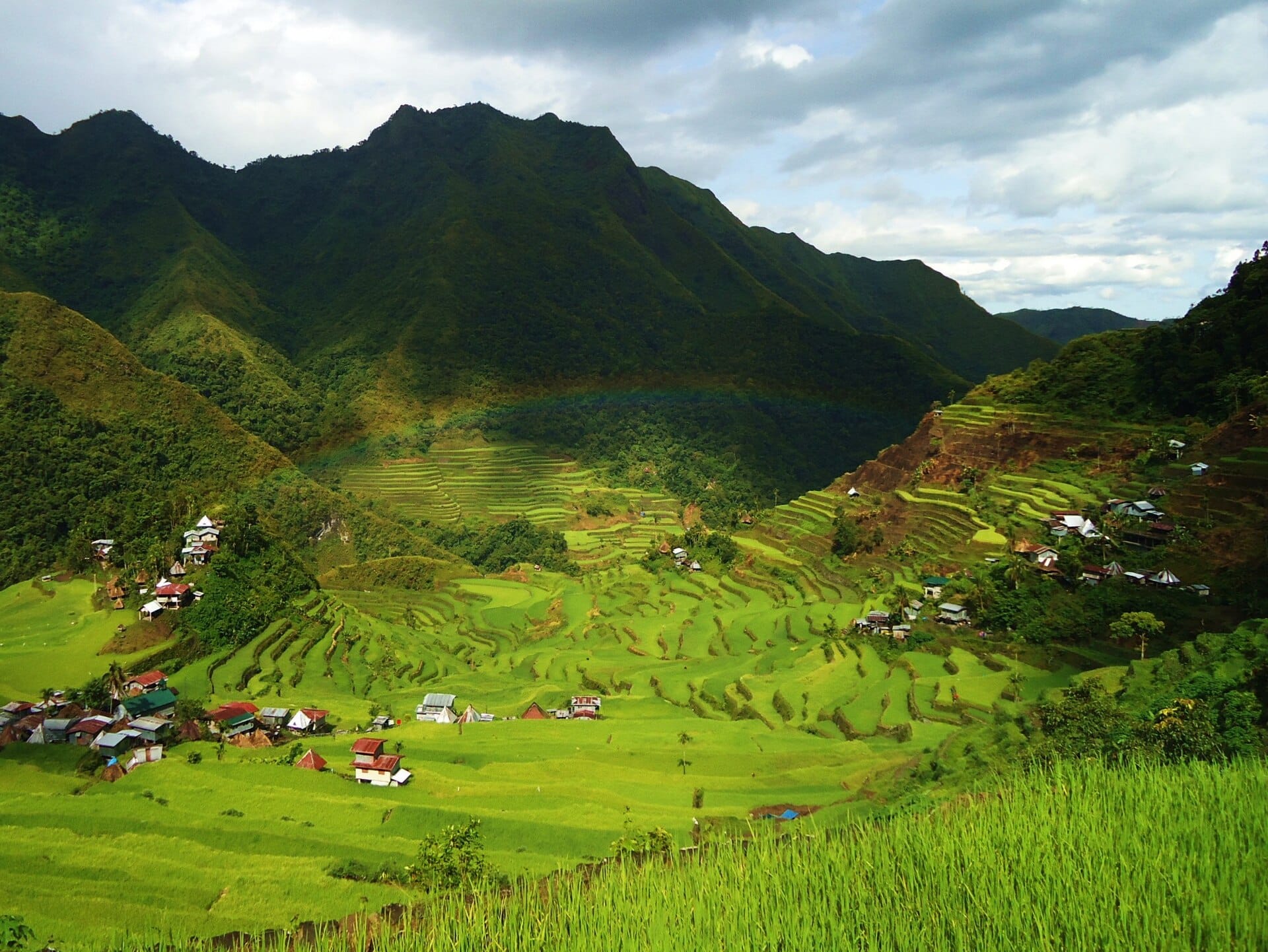 Faint rainbow over bright green rice terraces and village rooftops in a mountain valley at Batad