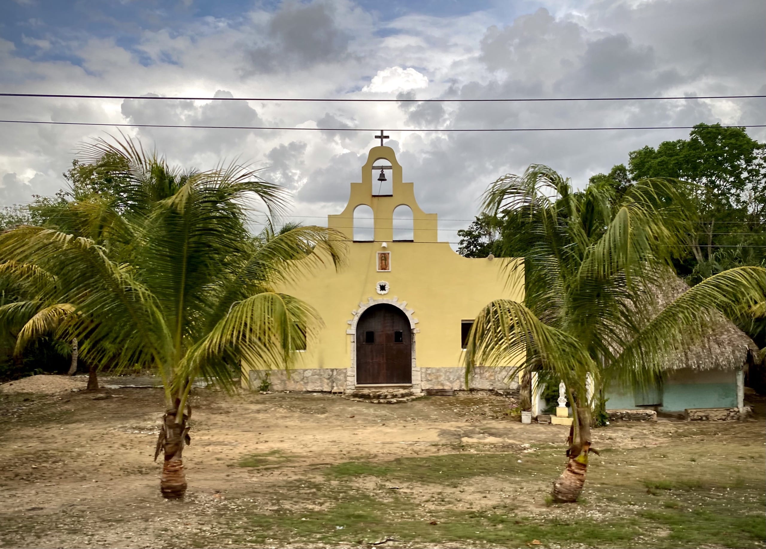 Yellow village church with a bell gable between two palms beside Highway 307 in Quintana Roo