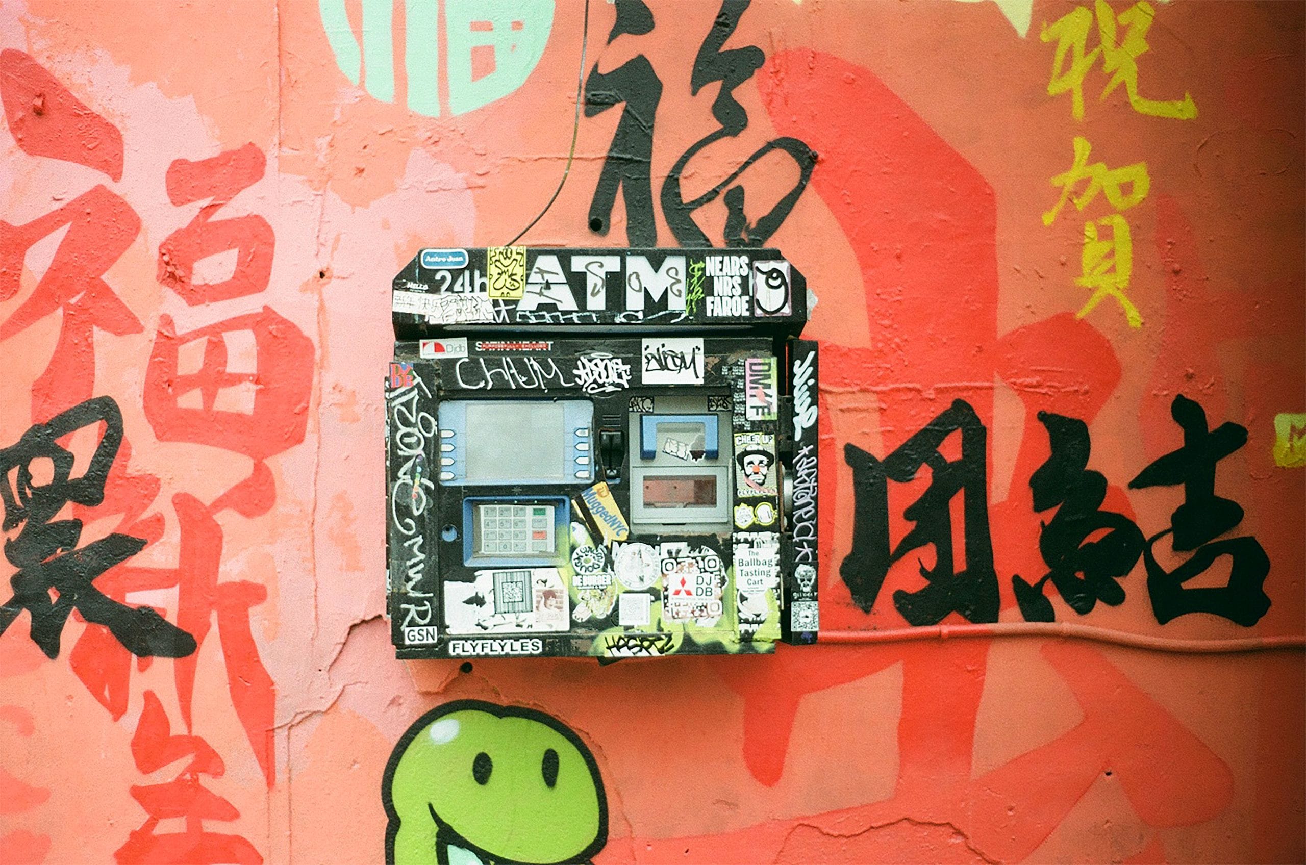 A black ATM covered in stickers mounted on a pink wall painted with large Chinese characters and graffiti