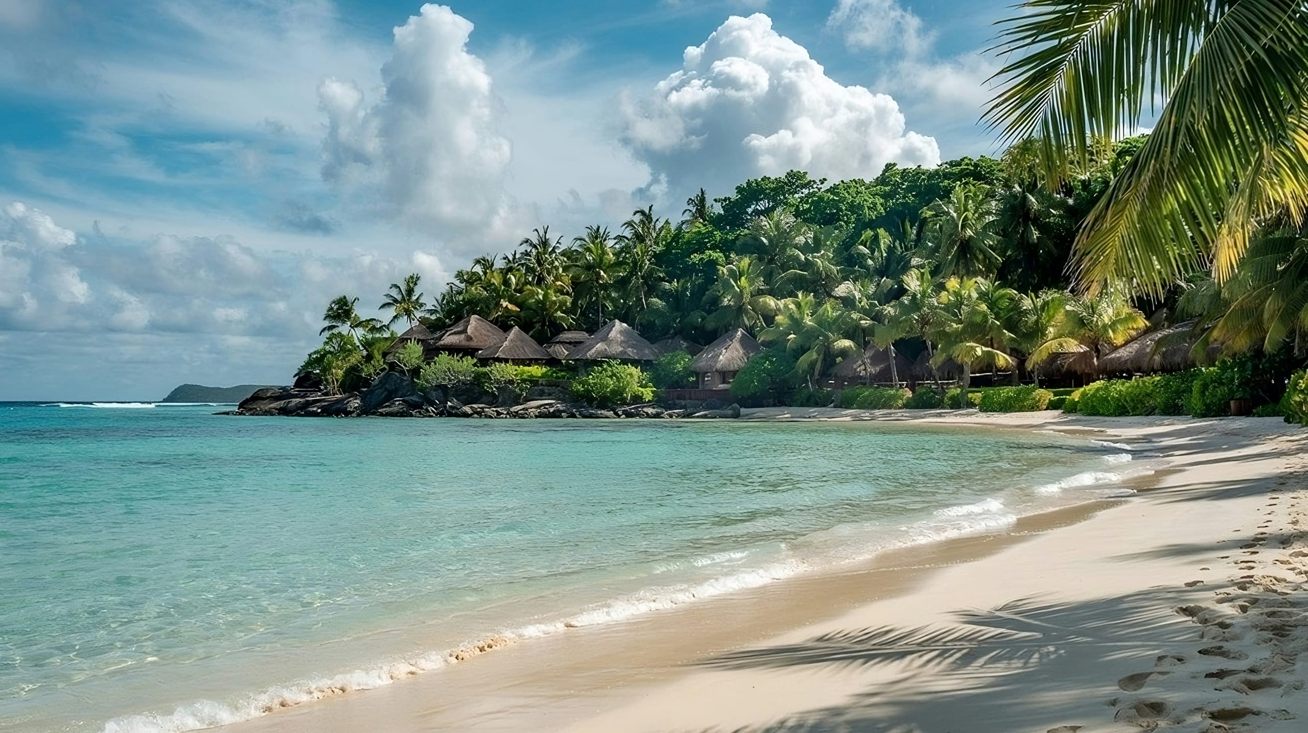 A white sand beach curving past thatched roofs under palms, footprints in the sand and pale turquoise water alongside