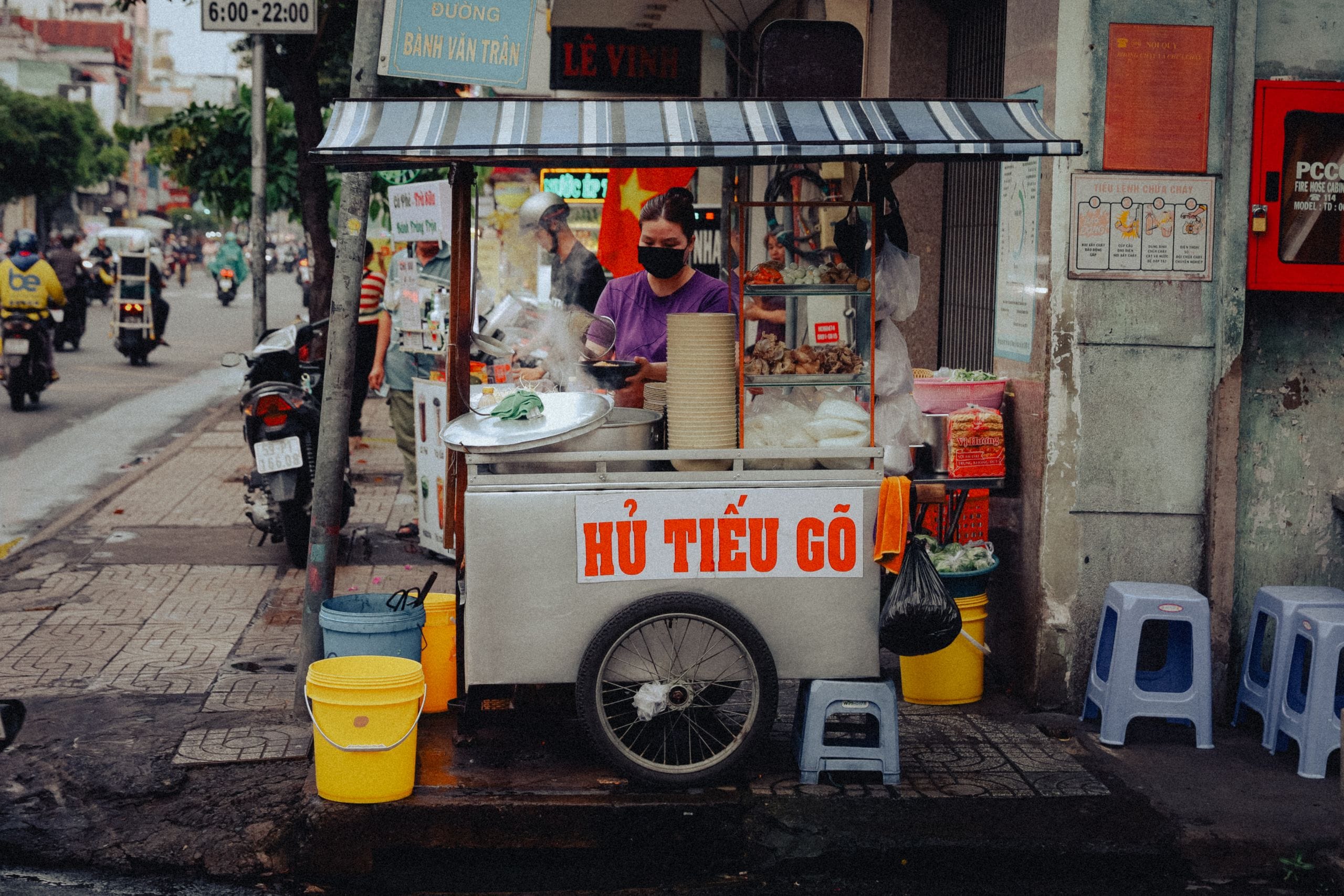 A noodle soup cart on a Ho Chi Minh City sidewalk, a woman serving behind it and plastic stools nearby