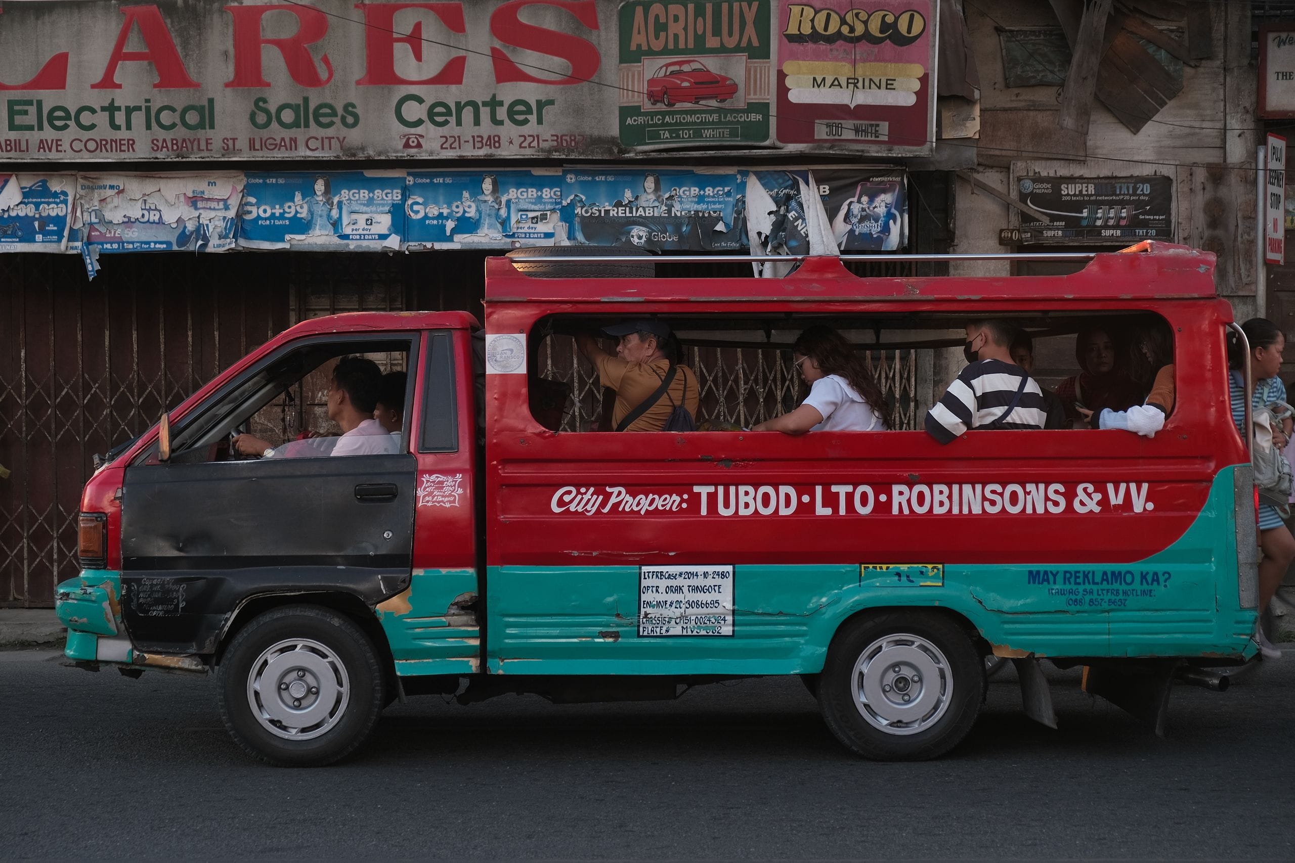A red and teal shared minibus on a city street, passengers sitting along the open windows behind the driver