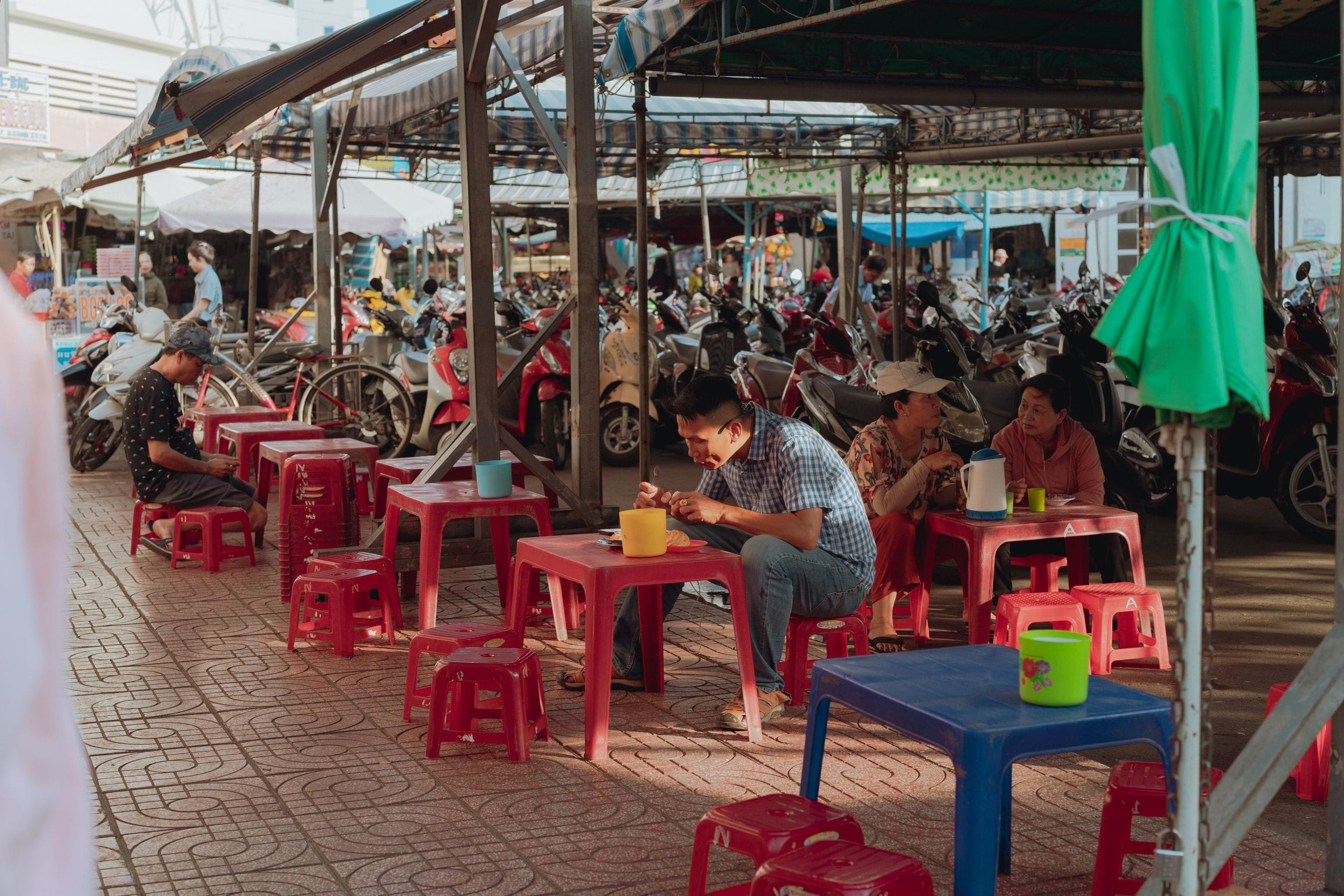 People eating at red and blue plastic tables under a market awning in Vietnam, motorbikes parked behind them
