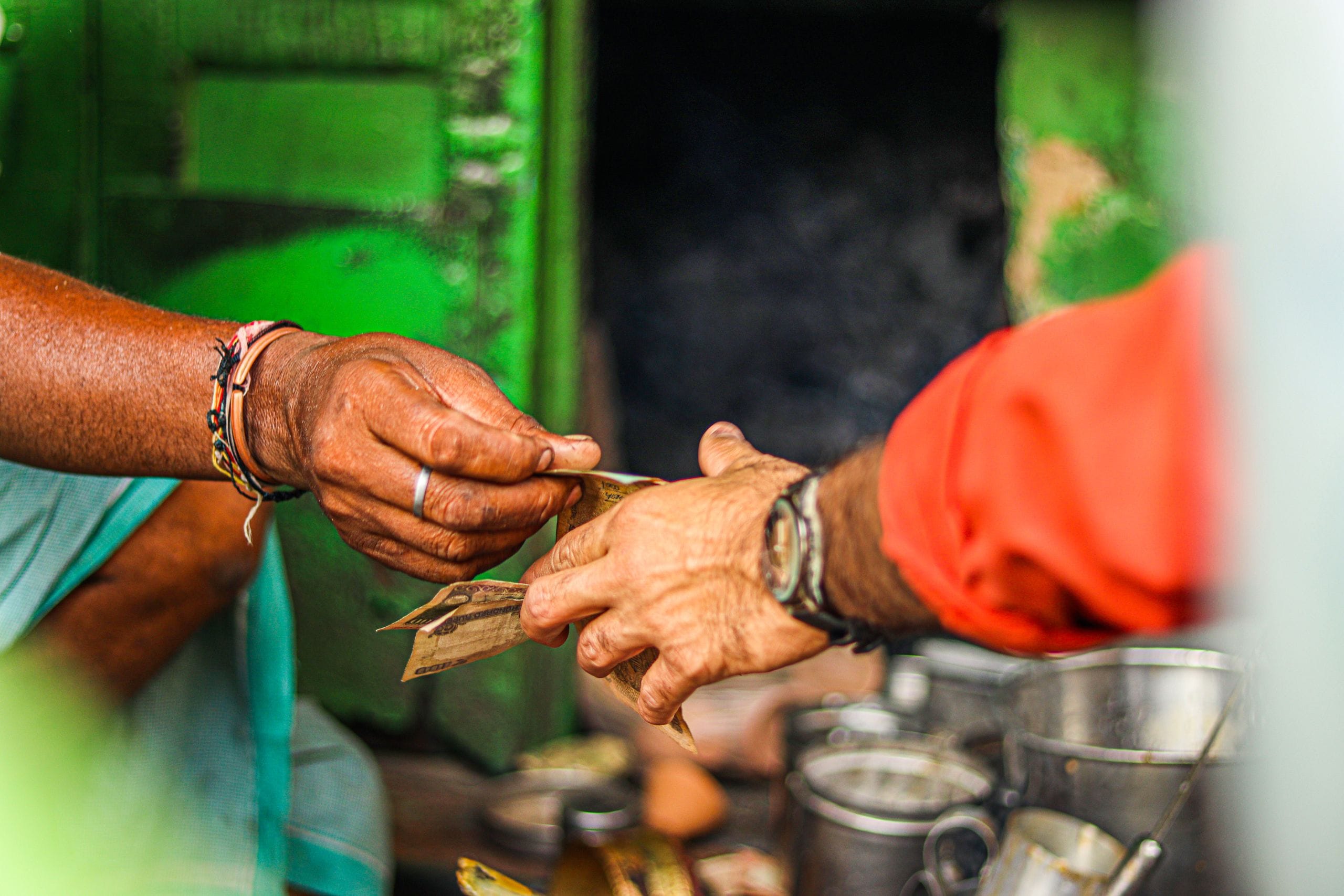Two hands passing folded paper banknotes across a street stall counter, steel cups in the foreground