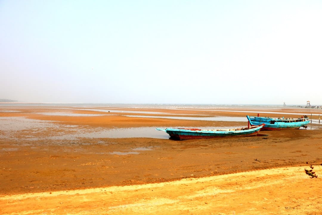 Two wooden boats aground on a wide tidal flat at low tide, the water gone out to a thin line in the haze