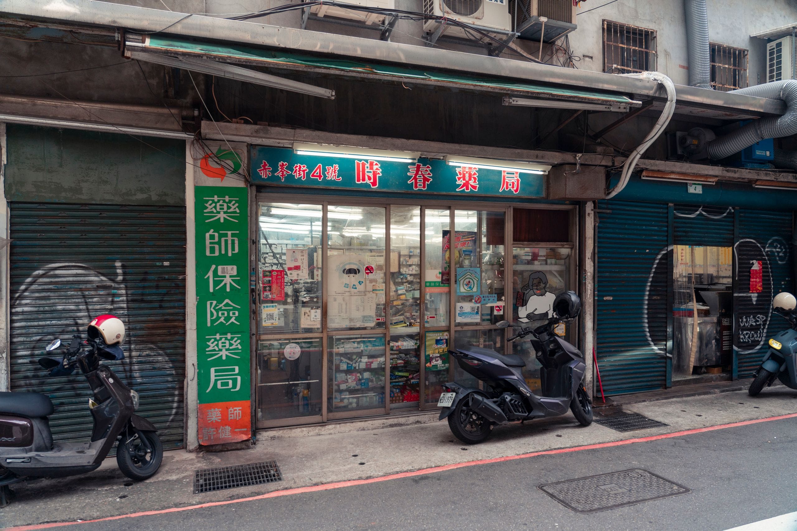 A narrow street pharmacy with Chinese lettering on its green signs and scooters parked by the door