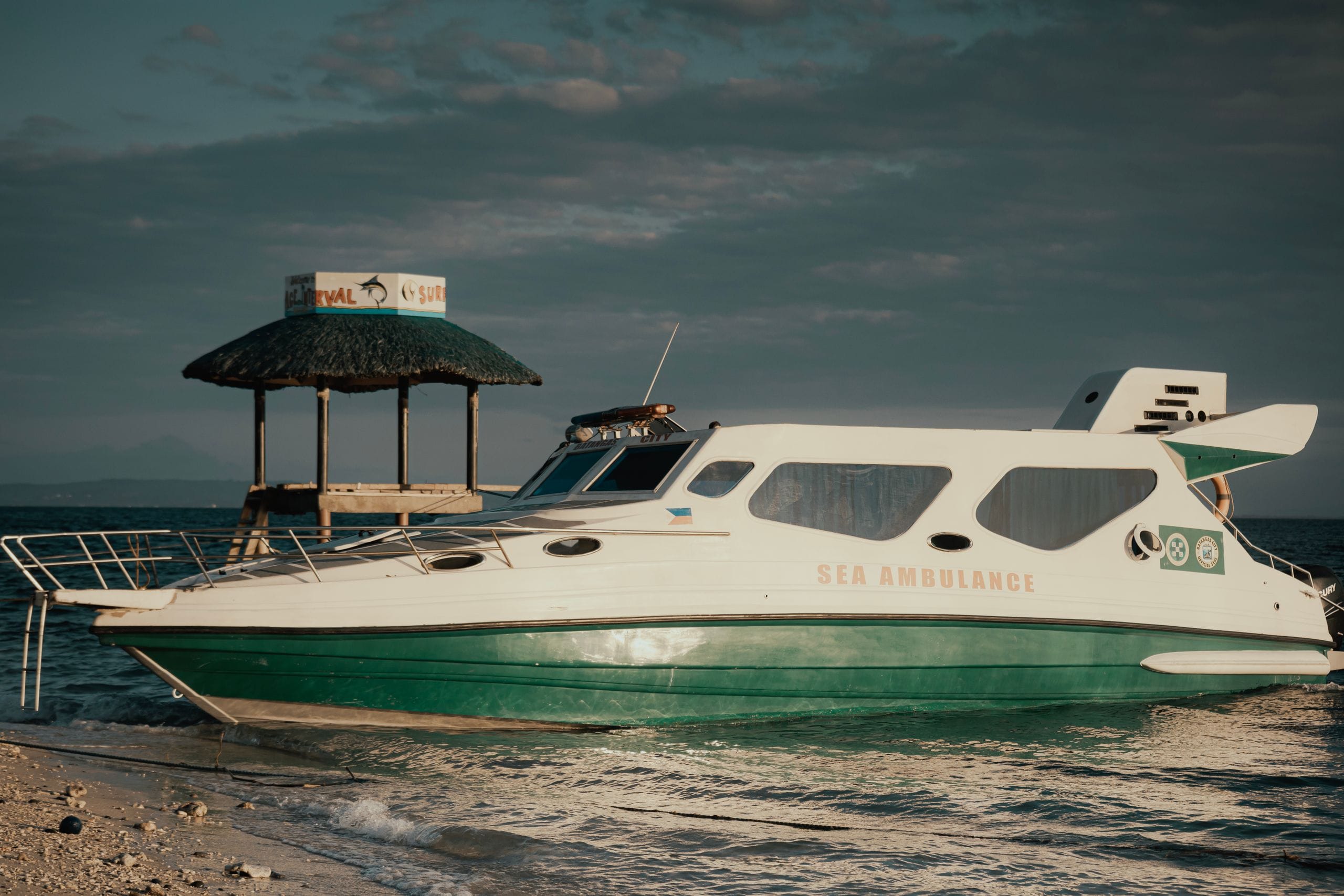 A white and green boat marked Sea Ambulance pulled up on a shallow beach beside a thatched shelter at dusk