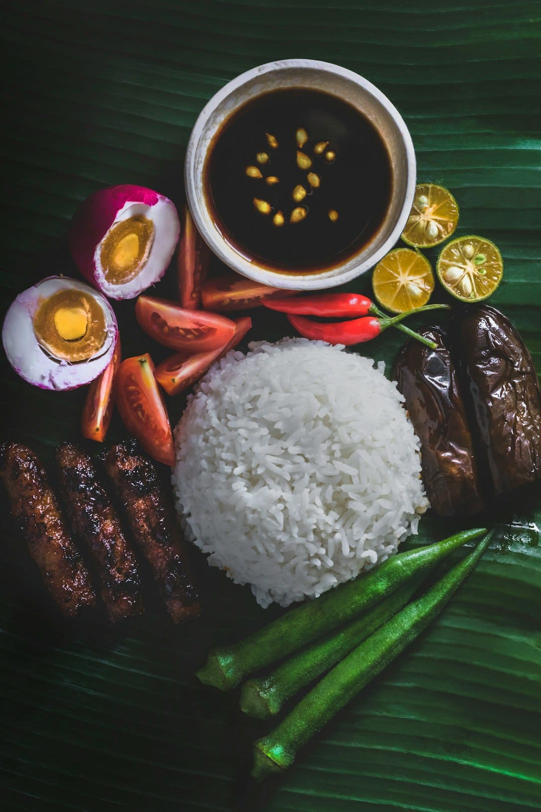 A banana leaf laid with white rice, grilled sausage, halved salted eggs, tomato, eggplant, okra and calamansi