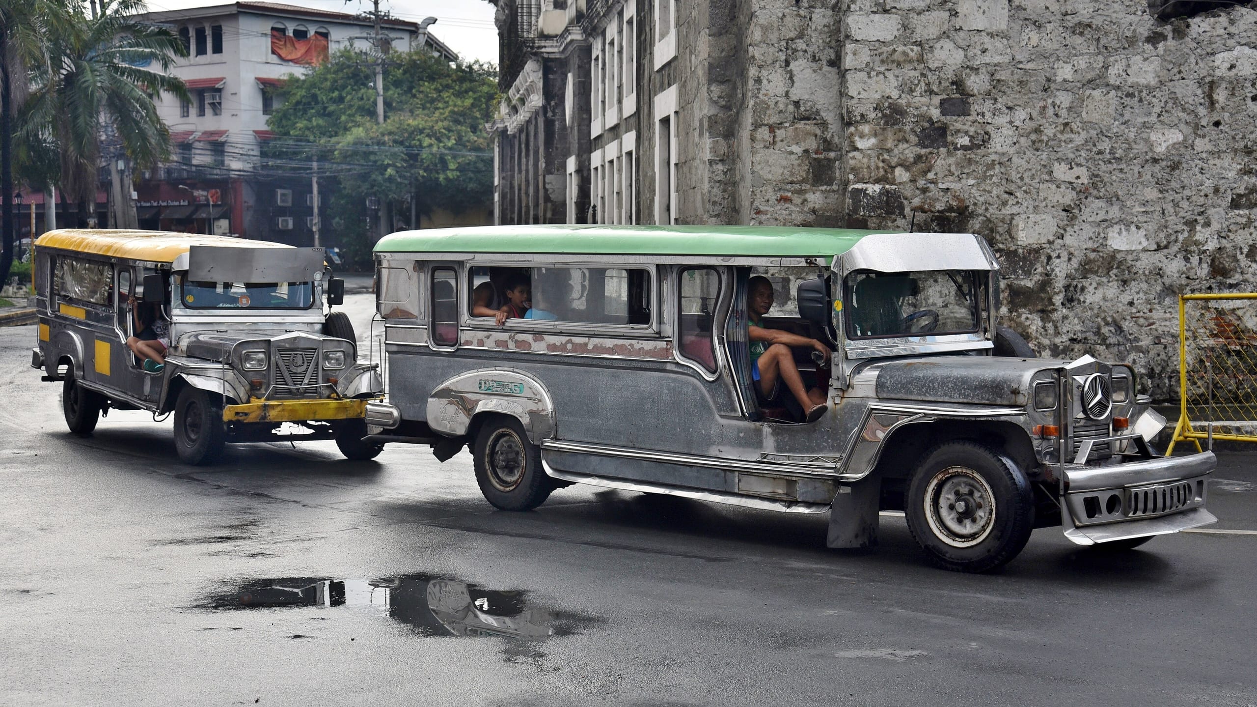 Two silver jeepneys turn along a wet street beside an old stone wall in Intramuros, Manila