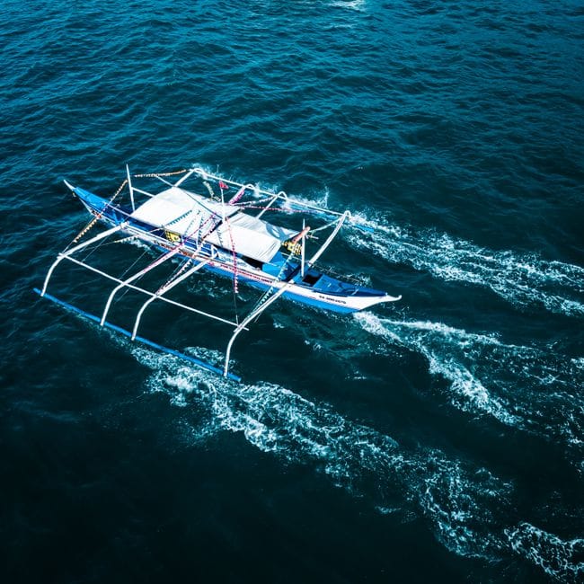 A single outrigger boat under way seen from above, its bamboo arms low over dark water and a wake trailing behind