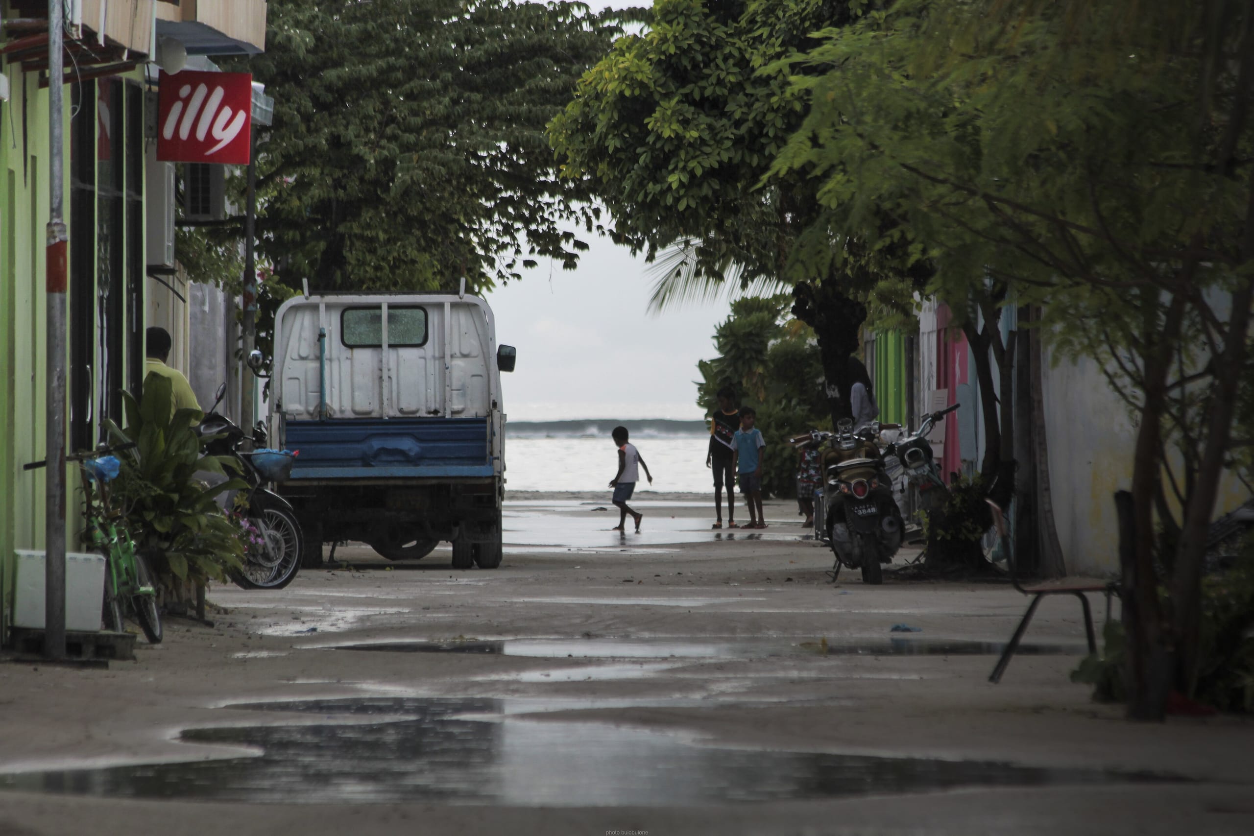 A sand street on Maafushi after rain, children running toward the sea at the far end