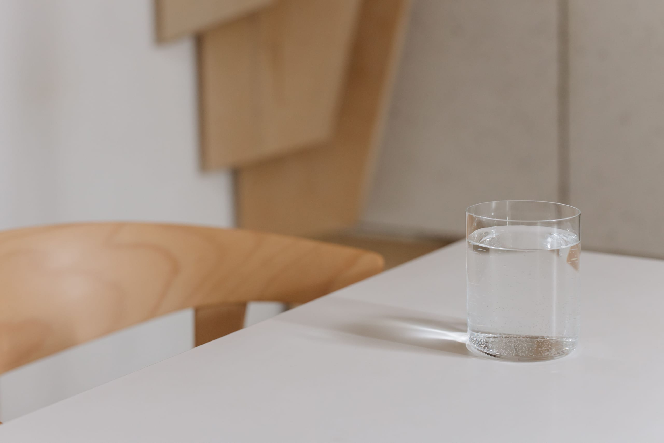 A plain glass of water on a white table beside a wooden chair