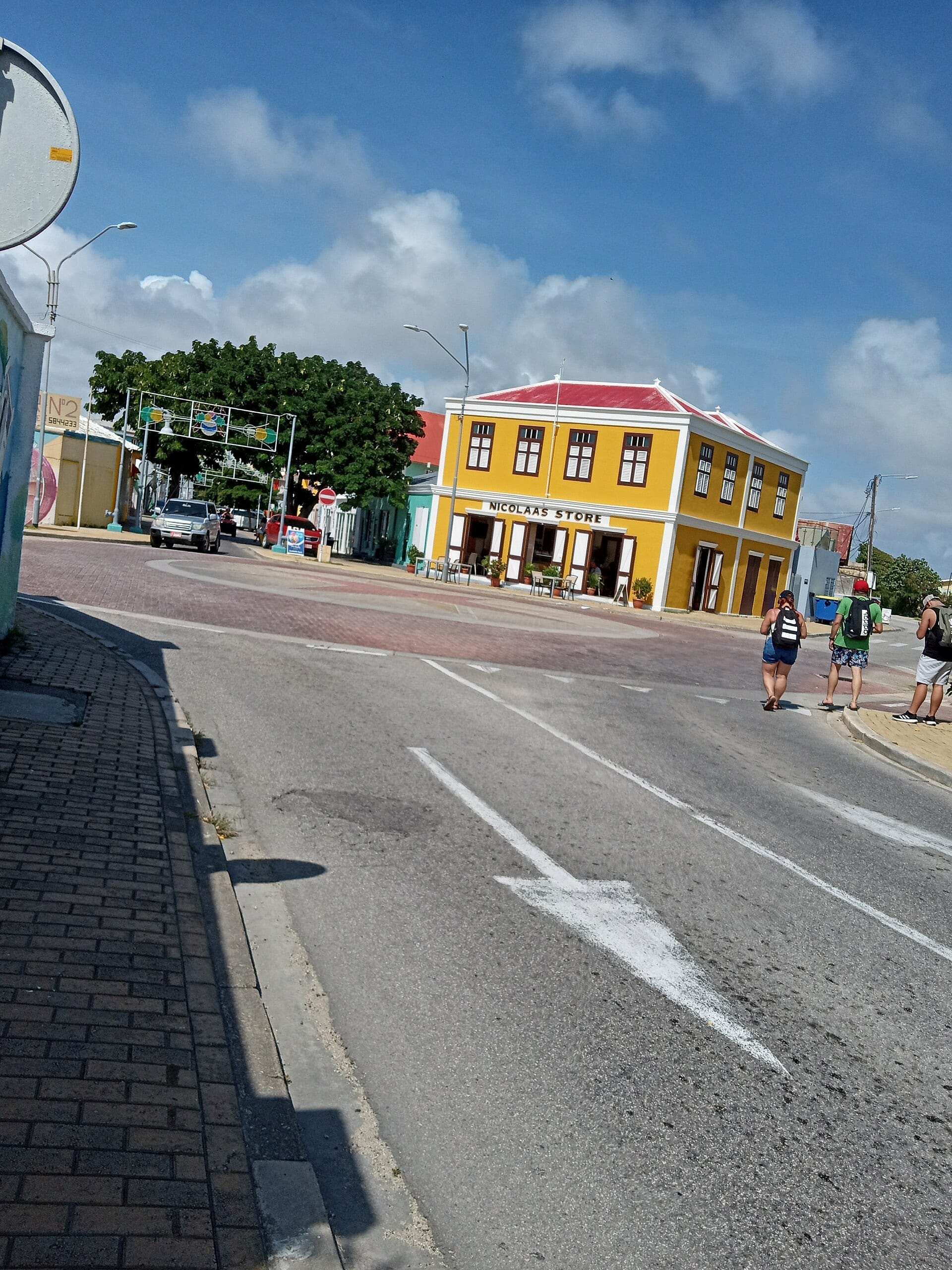 A wide road through a low Aruban town with a yellow colonial building on the corner and people waiting to cross