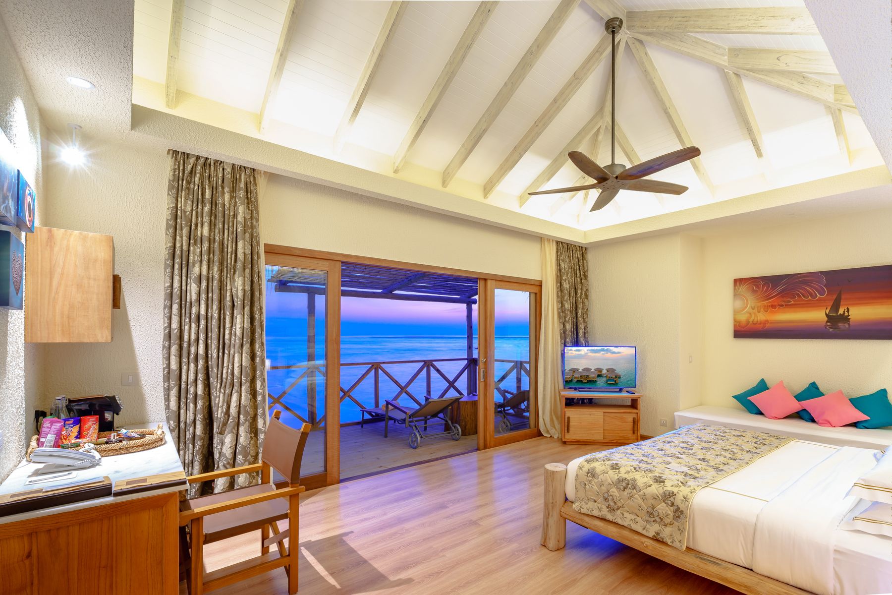 A resort room with a vaulted timber ceiling and a ceiling fan, sliding doors open onto a deck above the water