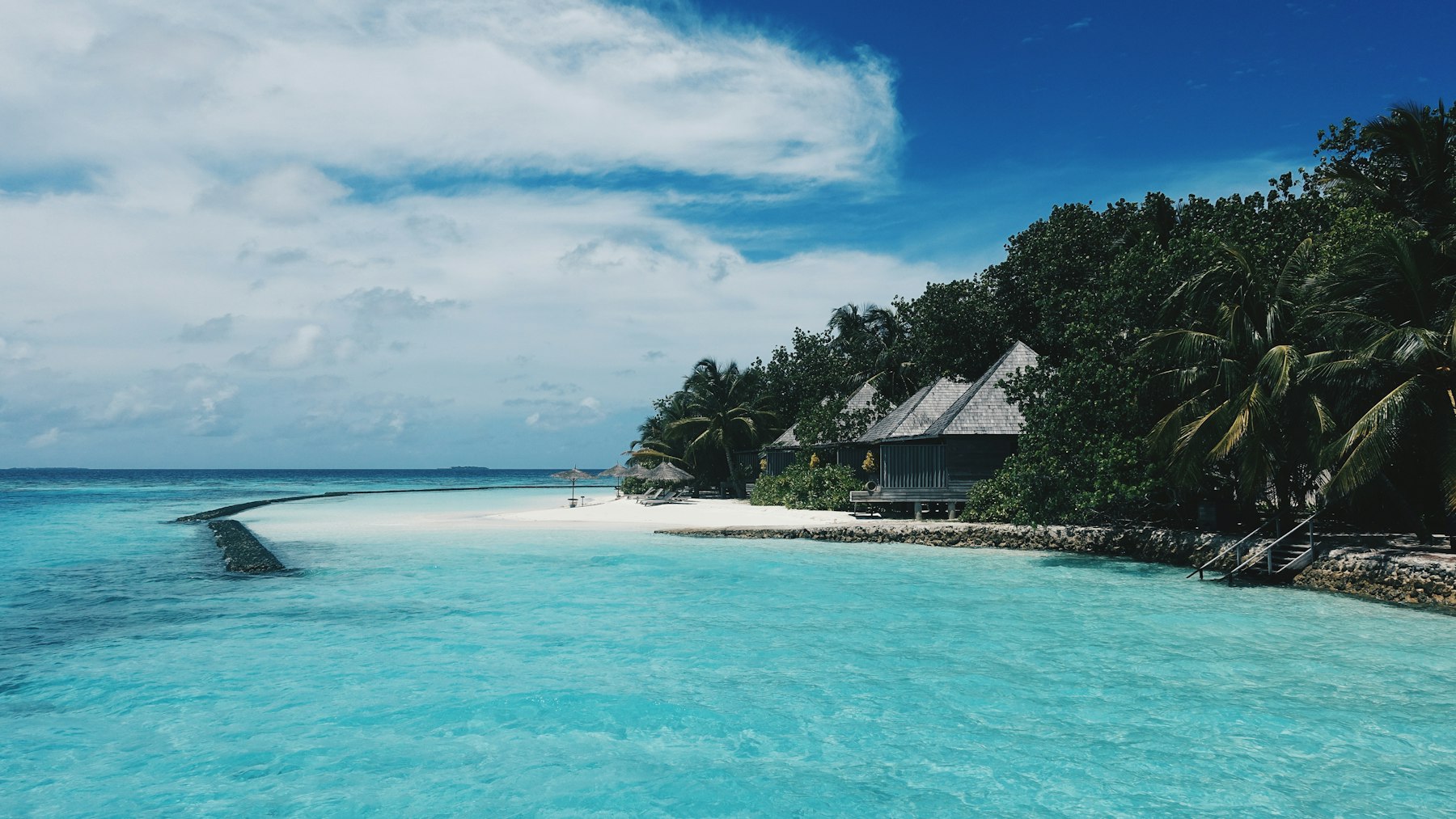 Beach villas under the trees at the edge of a shallow turquoise lagoon, sand running out to a low breakwater