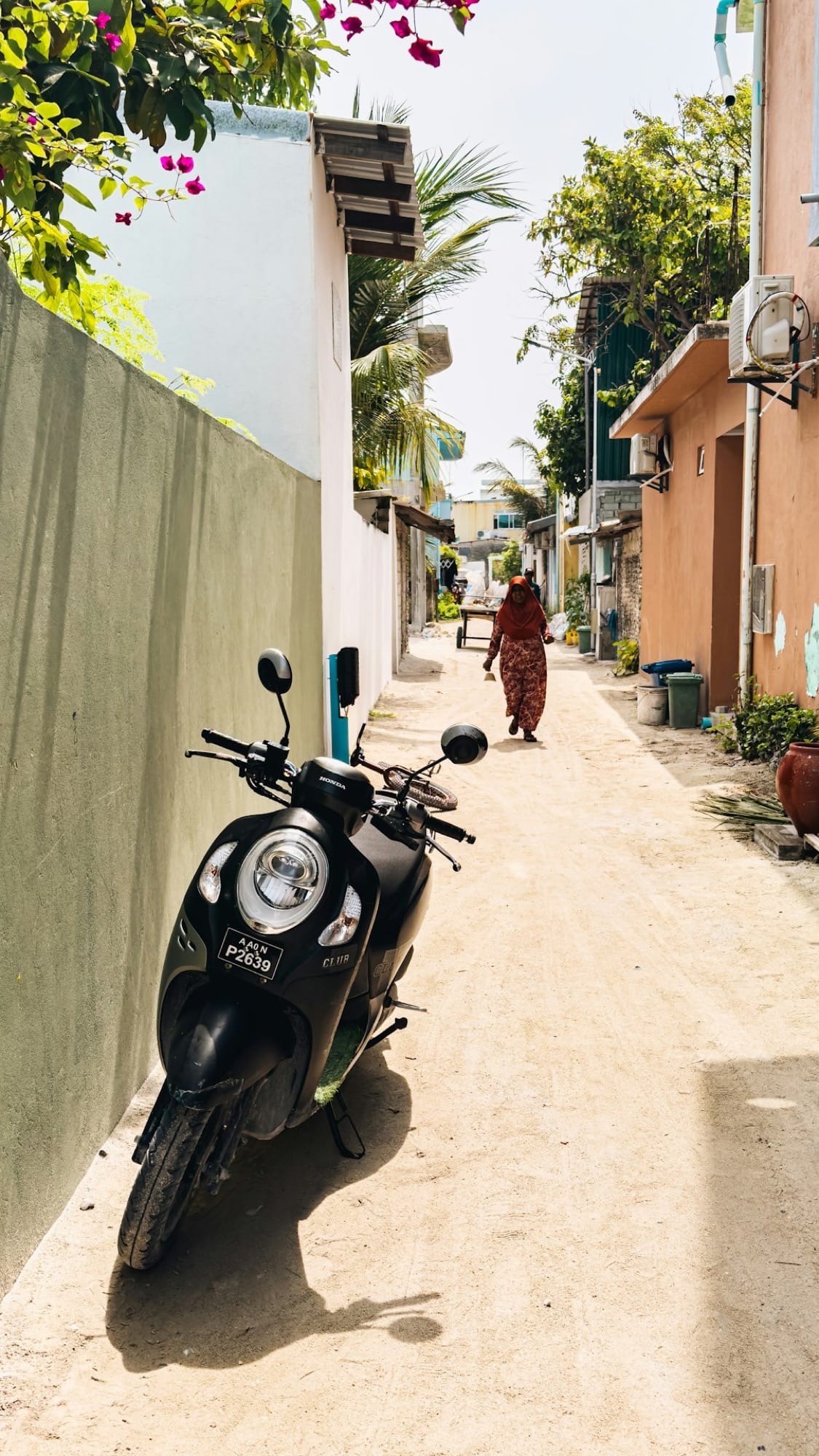 A sand lane between painted walls on an inhabited island, a scooter parked in the shade and a woman walking away from the camera